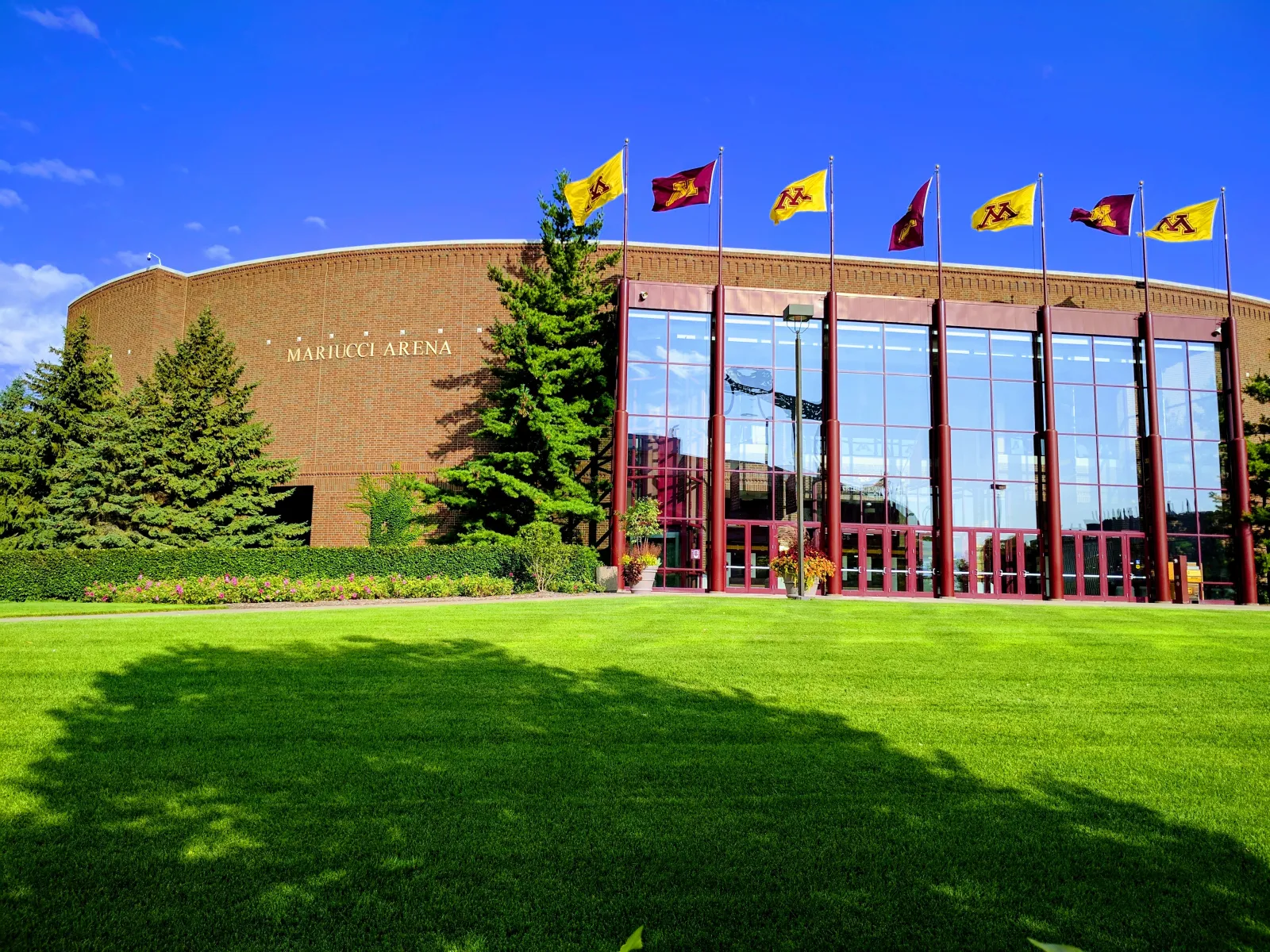 La entrada principal con frente de vidrio del 3M Arena at Mariucci, detrás del césped y las astas de bandera, en un edificio de ladrillo con un muro curvo. — 3M Arena at Mariucci