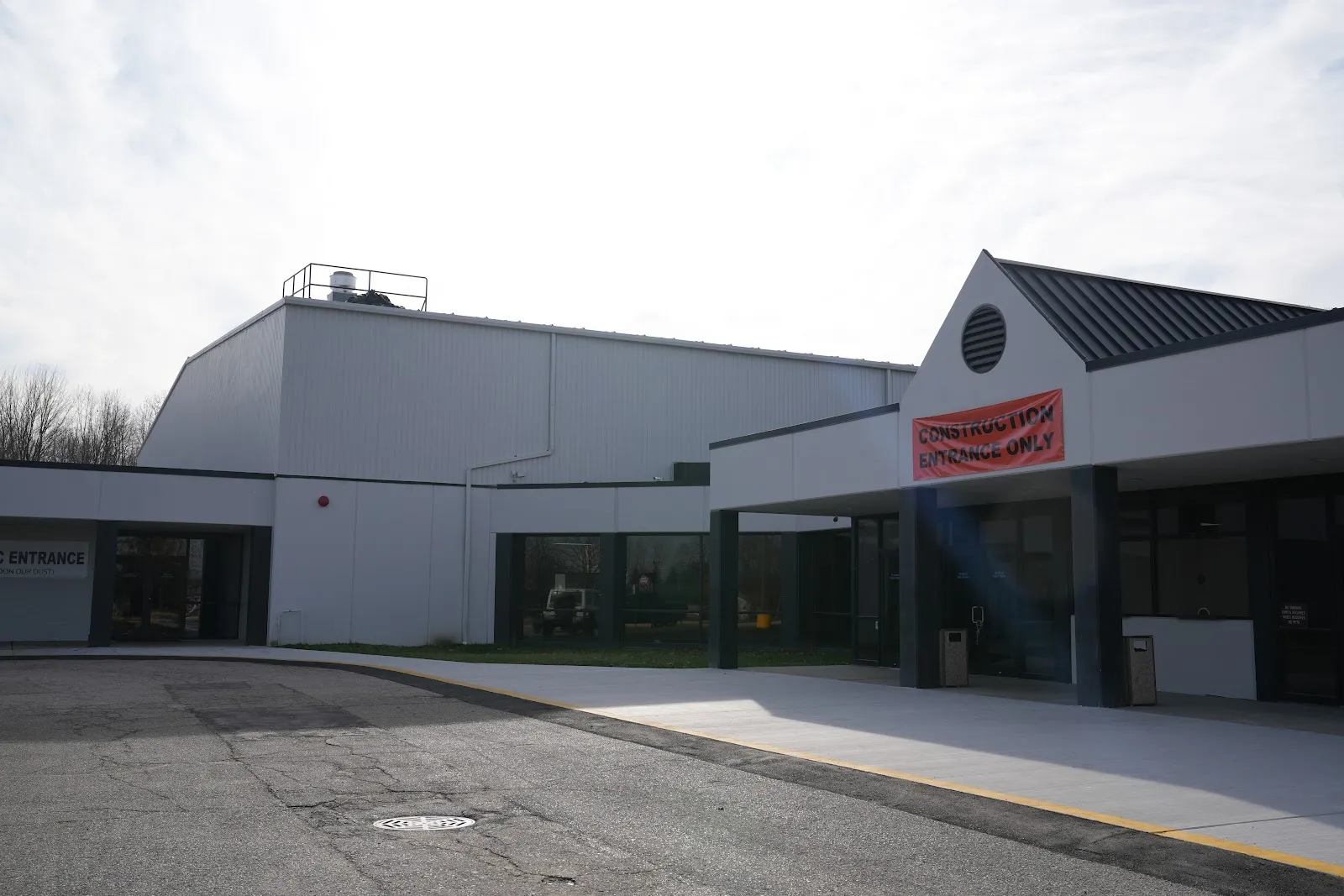 Front entrance of the 810 Sports Complex building on an overcast day, white metal-panel walls, a dark peaked-roof entry canopy over glass doors, and a temporary red "Construction Entrance Only" banner above one doorway. — 810 Sports Complex