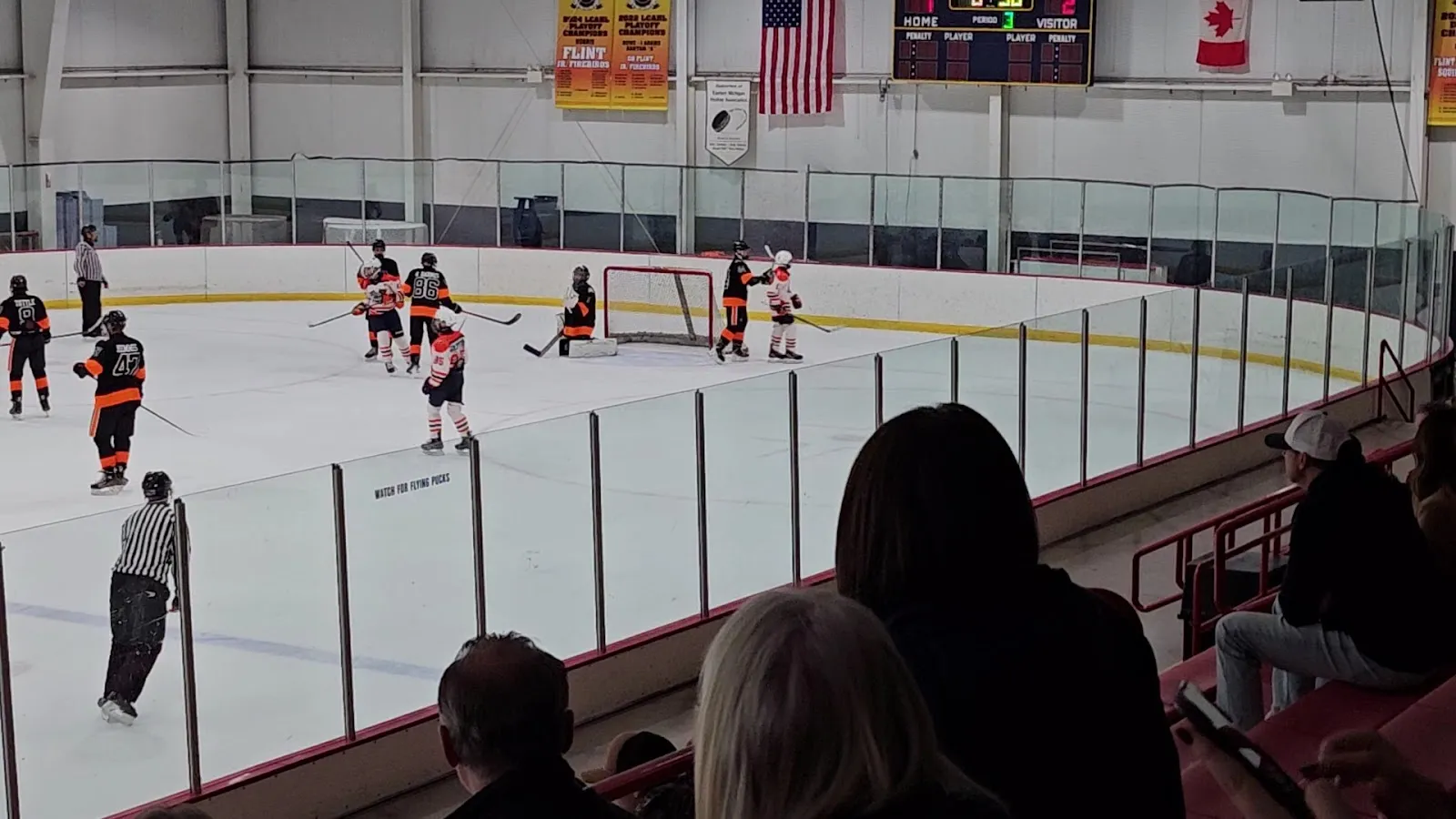 A hockey game on the indoor ice seen from the seating, players grouped around the far net under a scoreboard and flags, with spectators in the foreground. — 810 Sports Complex