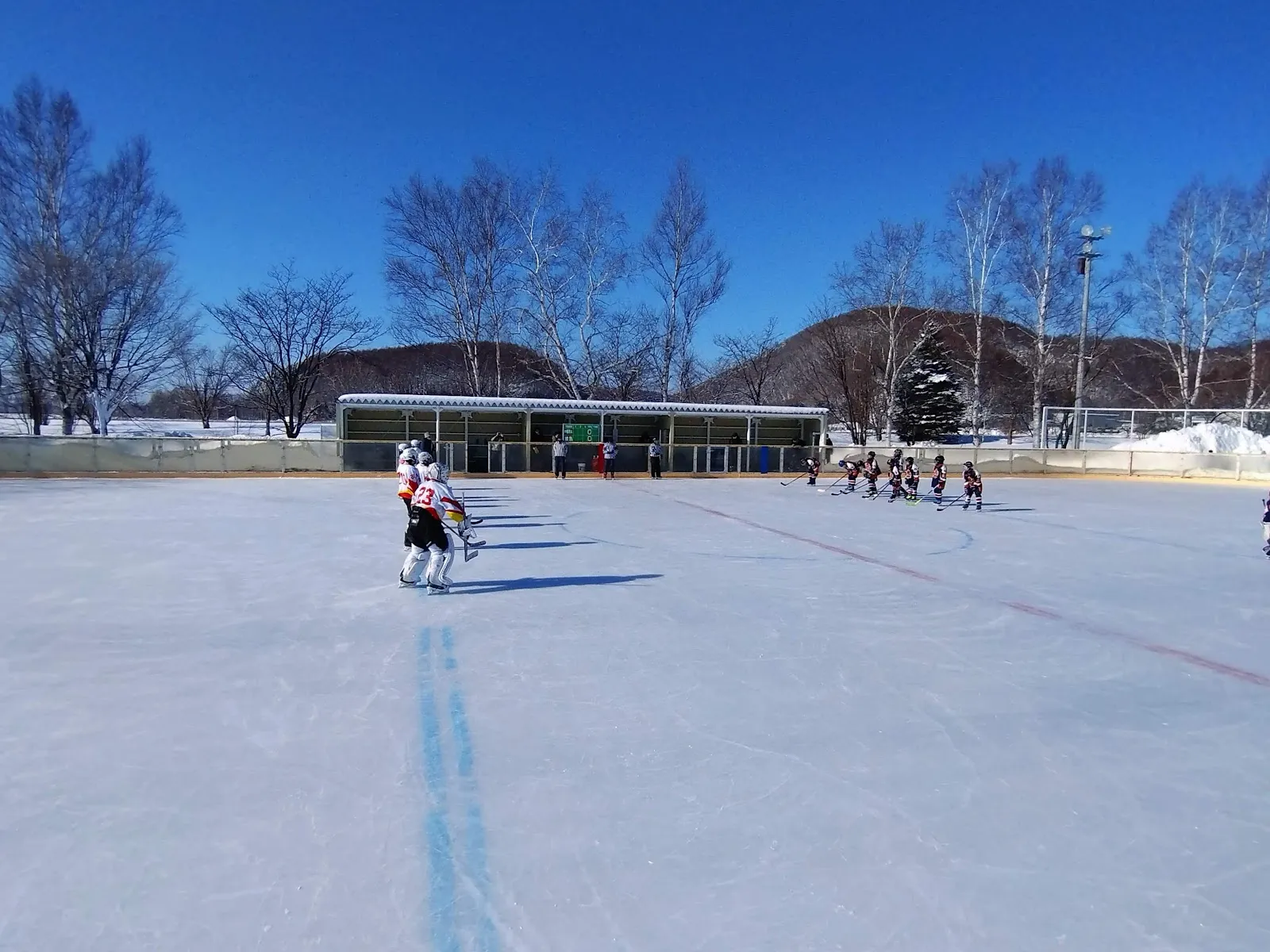 雪に縁取られた網走の天然氷リンク。遠くでは少年アイスホッケーの試合が行われ、フェンスの奥には屋根付きの休憩所が見えます。 — Abashiri City Skating Rink