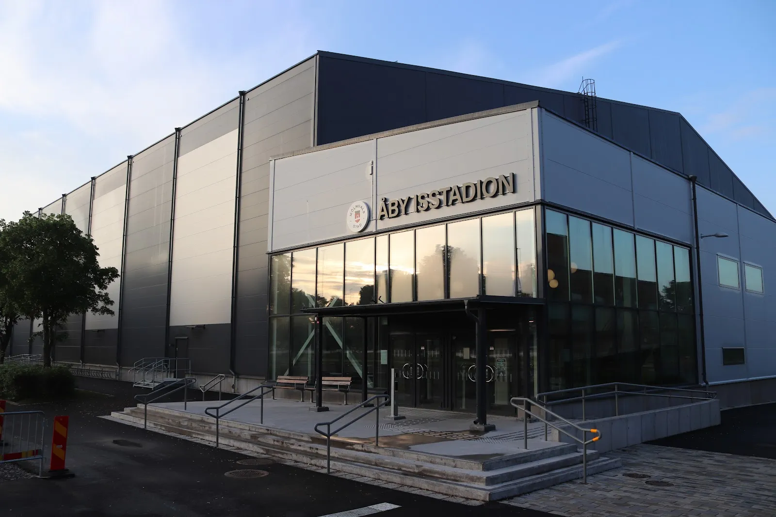 Entrésidan av Åby isstadion i Mölndal med namnet och Mölndals stads emblem ovanför en glasentré, trappor och bänkar, och den grå plåtklädda hallen bakom – Åby isstadion (Mölndal)