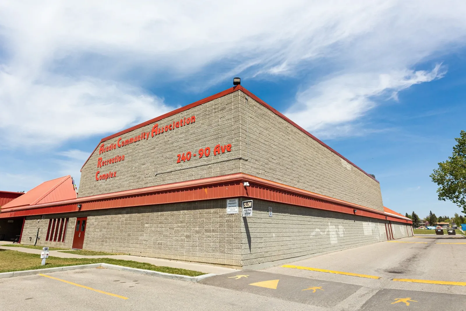 El costado de ladrillo y bloques del complejo bajo un cielo azul, con Acadia Community Association Recreation Complex y 240 - 90 Ave pintados en letras rojas en lo alto del muro. — Acadia Recreation Complex