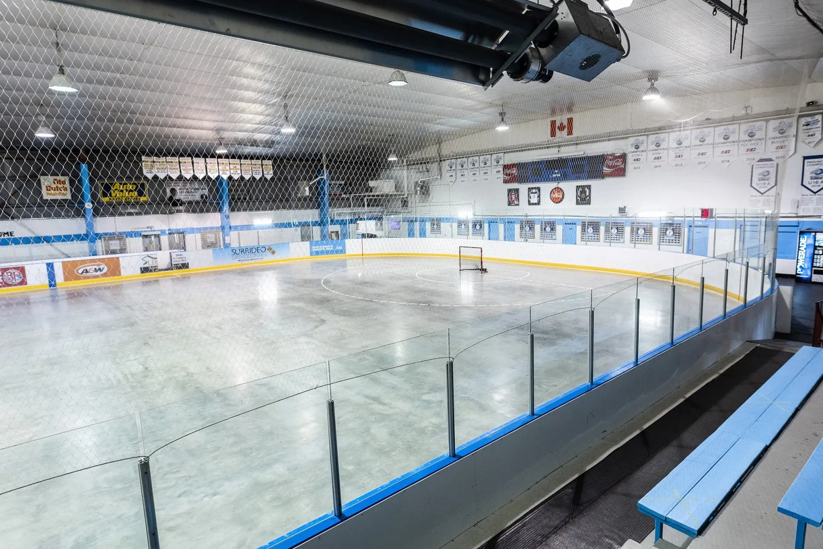 La arena vacía vista desde las bancas azules detrás del cristal, con mantas de campeonato y una bandera de Canadá a lo largo de la pared del fondo. — Acadia Recreation Complex