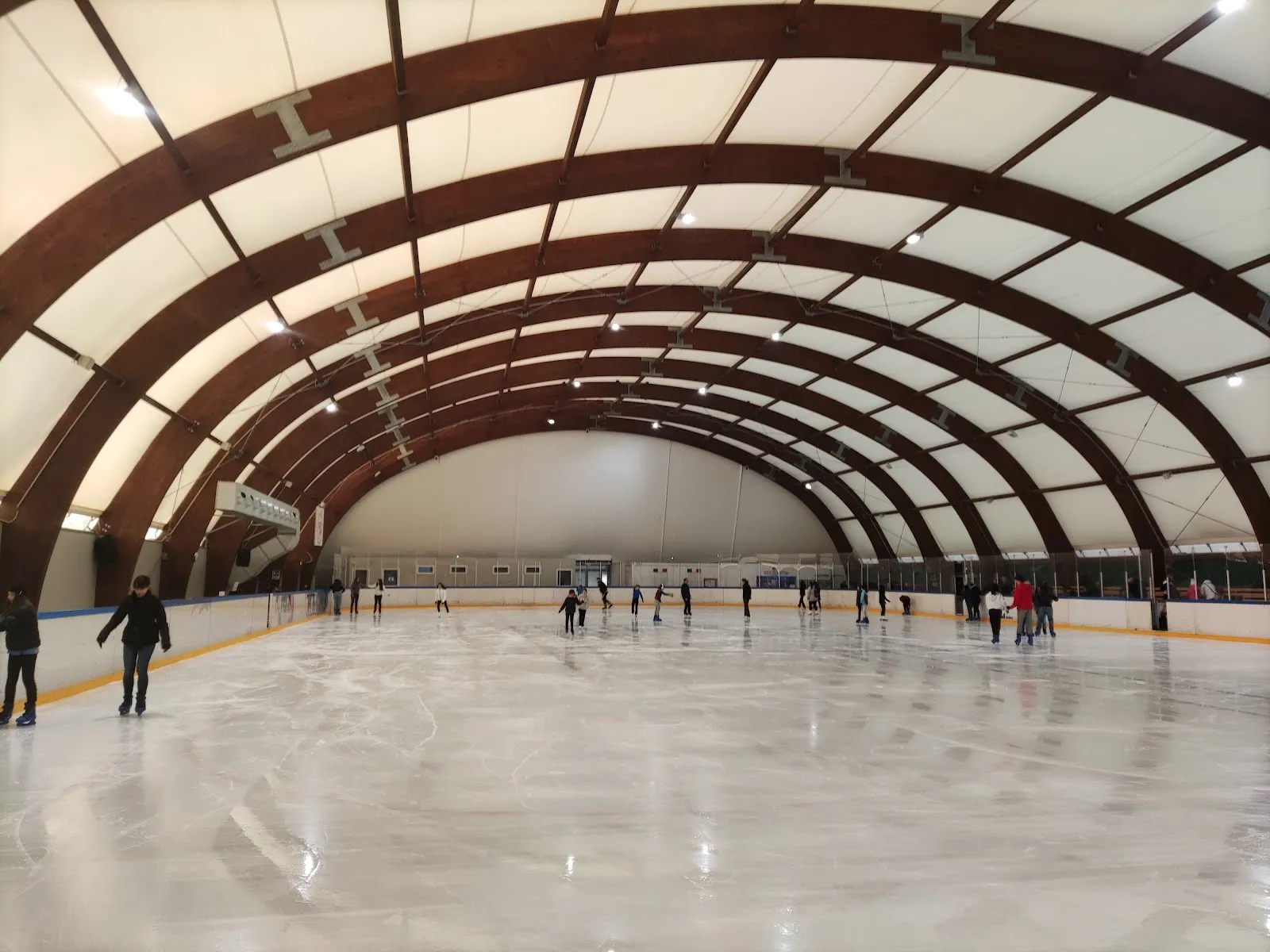 Wide view across the vaulted San Donato Milanese ice hall with brown timber arches and skaters spread along the far side. — Accademia del Ghiaccio