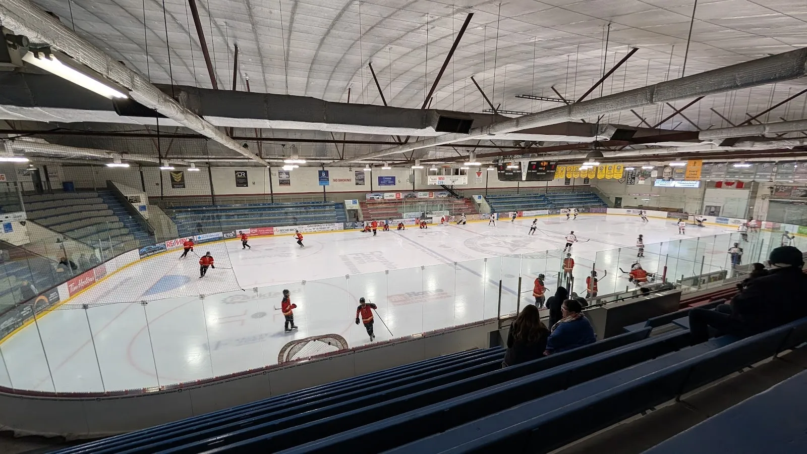 A game seen from the stands, players spread across a wide sheet under a steel-trussed roof with a banner row along the far wall. — Access Communications Centre