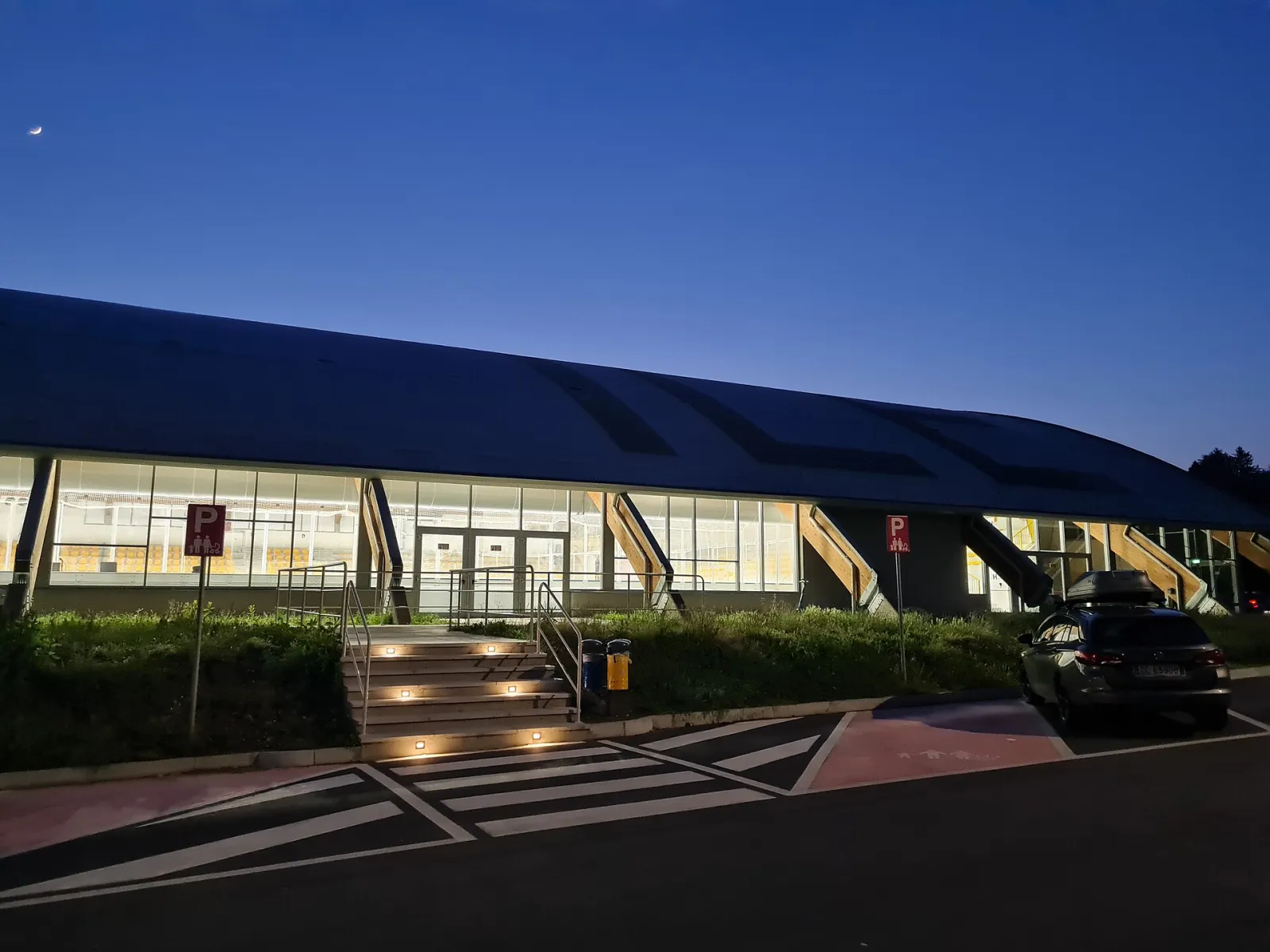 The long curved roof and glazed front of the Acinque Ice Arena in Varese at dusk, with the entrance steps and car park in front. — Acinque Ice Arena (PalAlbani)