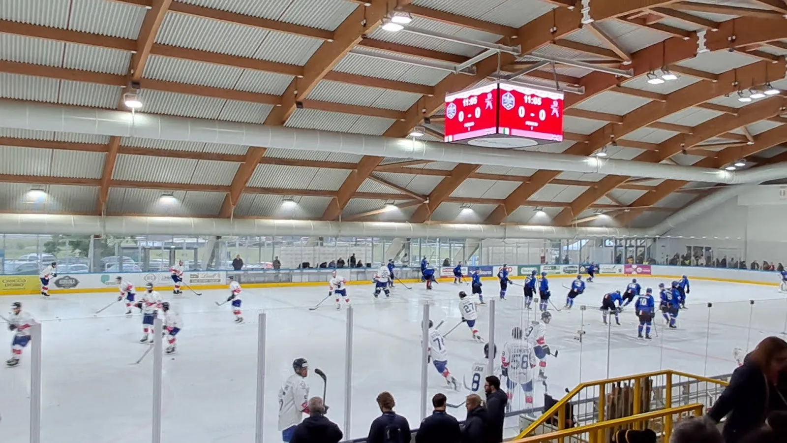 An ice hockey game under way in the Varese ice arena, players spread across the rink beneath the exposed timber roof beams with the scoreboard overhead. — Acinque Ice Arena (PalAlbani)