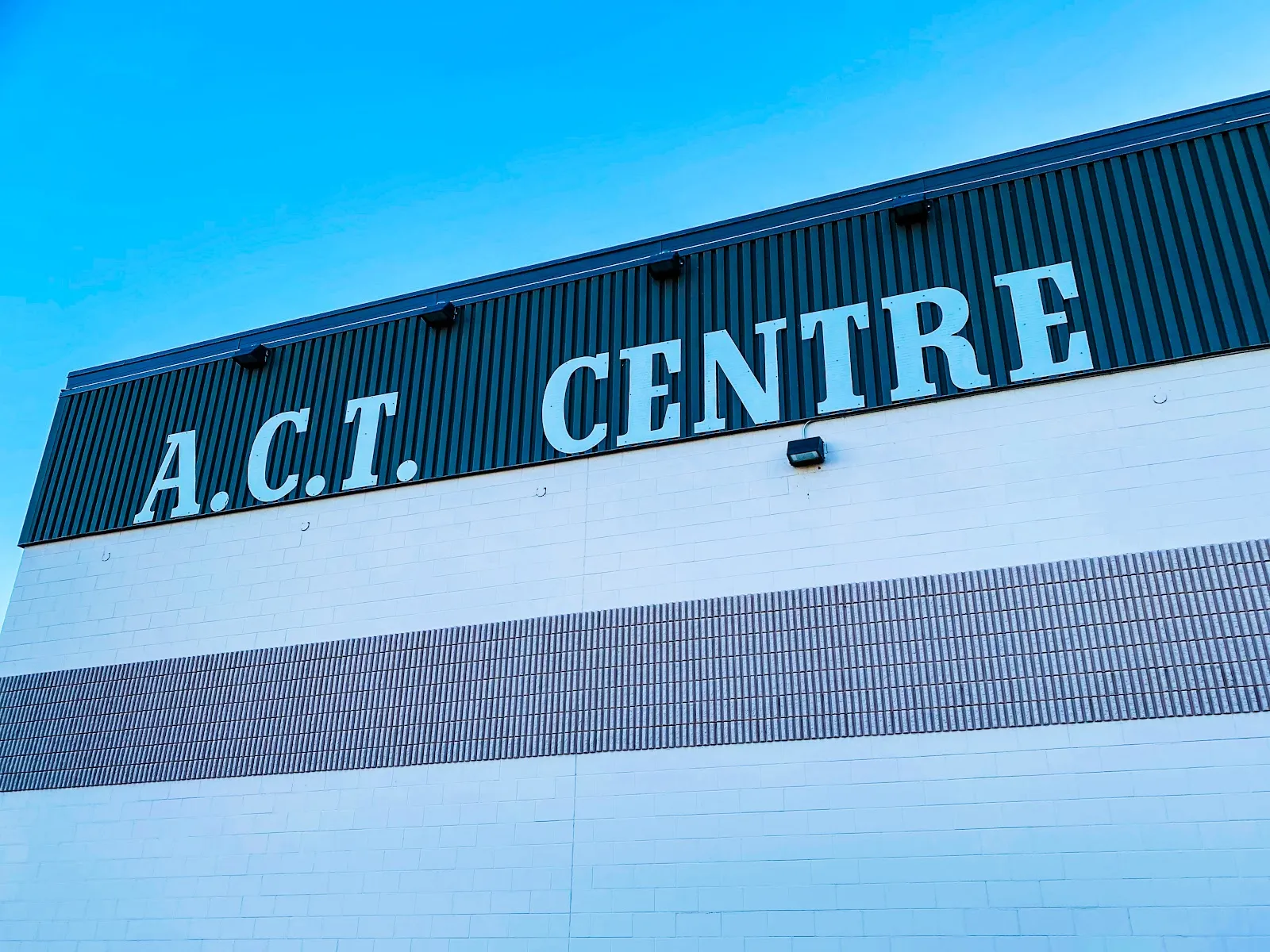 Low-angle view of the building's upper wall against a blue sky, with large 'A.C.T. CENTRE' lettering on the dark metal fascia above the white block wall. — ACT Arena
