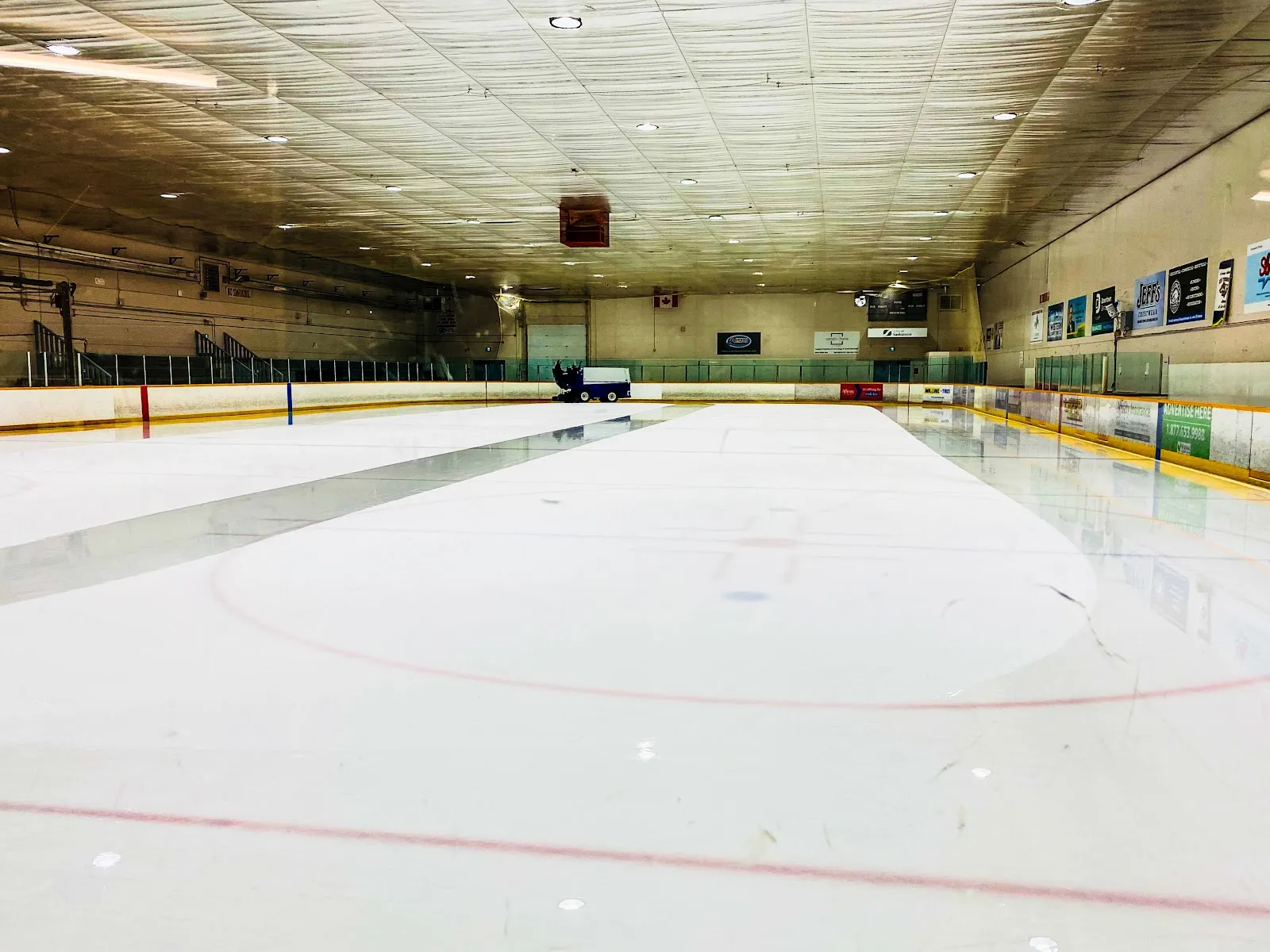 Empty full ice sheet freshly resurfaced, viewed from the stands, with the Zamboni parked at center ice, a Canadian flag, sponsor boards, and a 'City of Saskatoon' rink-board ad. — ACT Arena