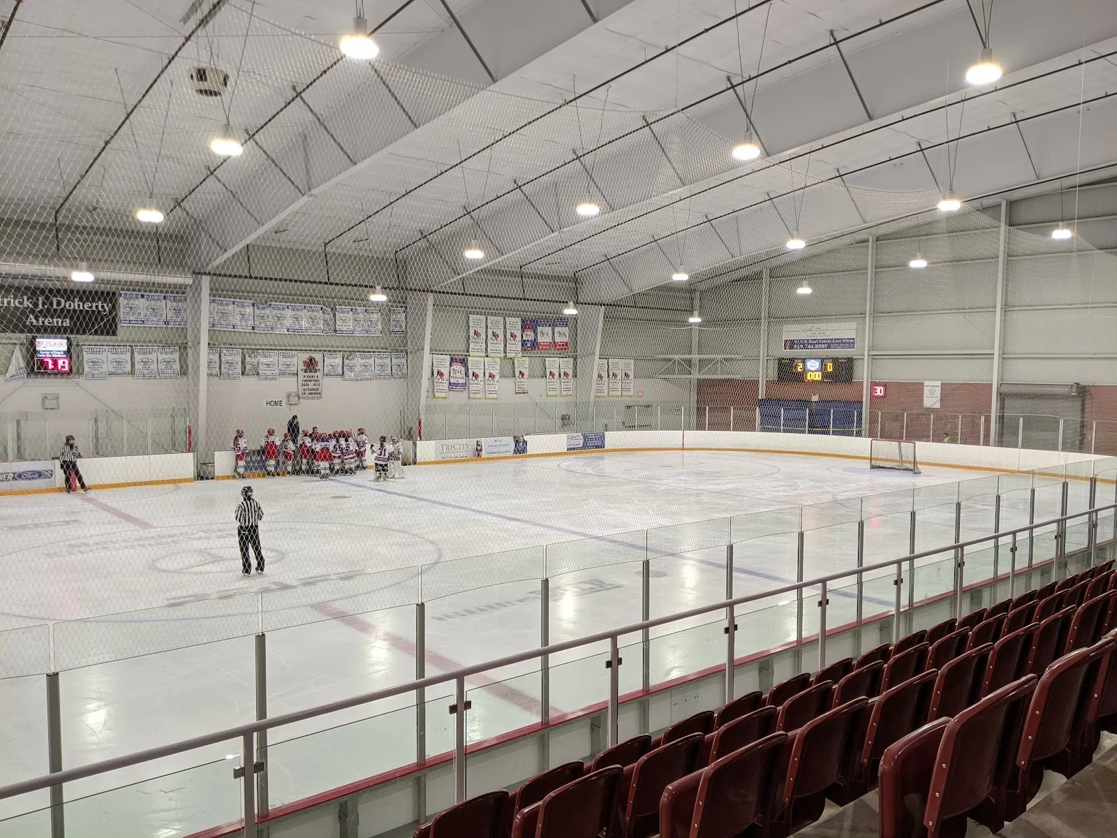 A wide empty sheet of ice seen from beside the red seats, a few skaters at the far end under a high white roof with Canadian flags on the end wall. — Activa Sportsplex