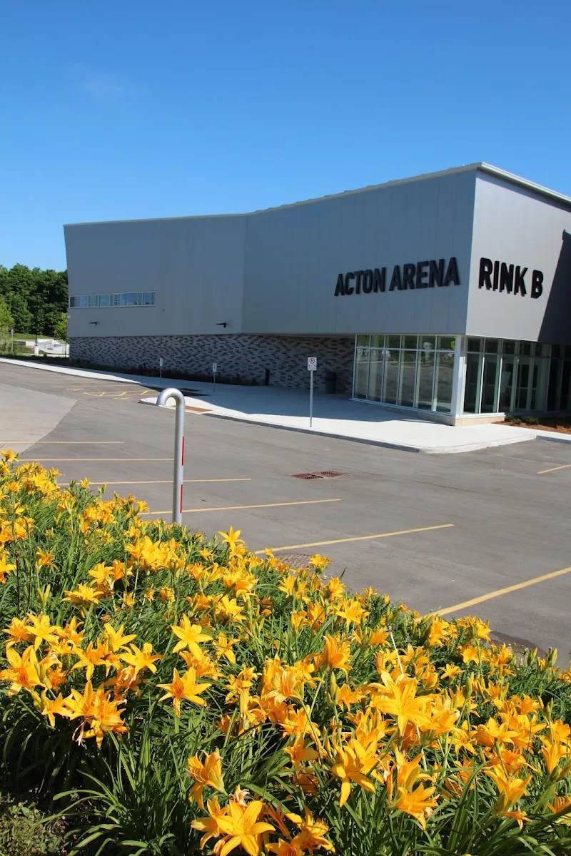 Acton Arena, revestida de metal gris, en un día soleado, con "Acton Arena Rink B" en la pared, la entrada de cristal y un macizo de lirios naranjas al frente. — Acton Arena and Community Centre