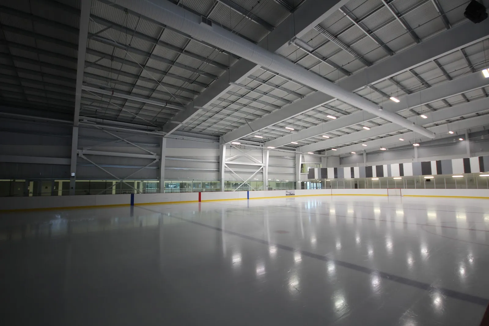 Una pista vacía de Acton Arena bajo el techo blanco de acero, con el cristal y las vallas a lo largo del lado opuesto. — Acton Arena and Community Centre