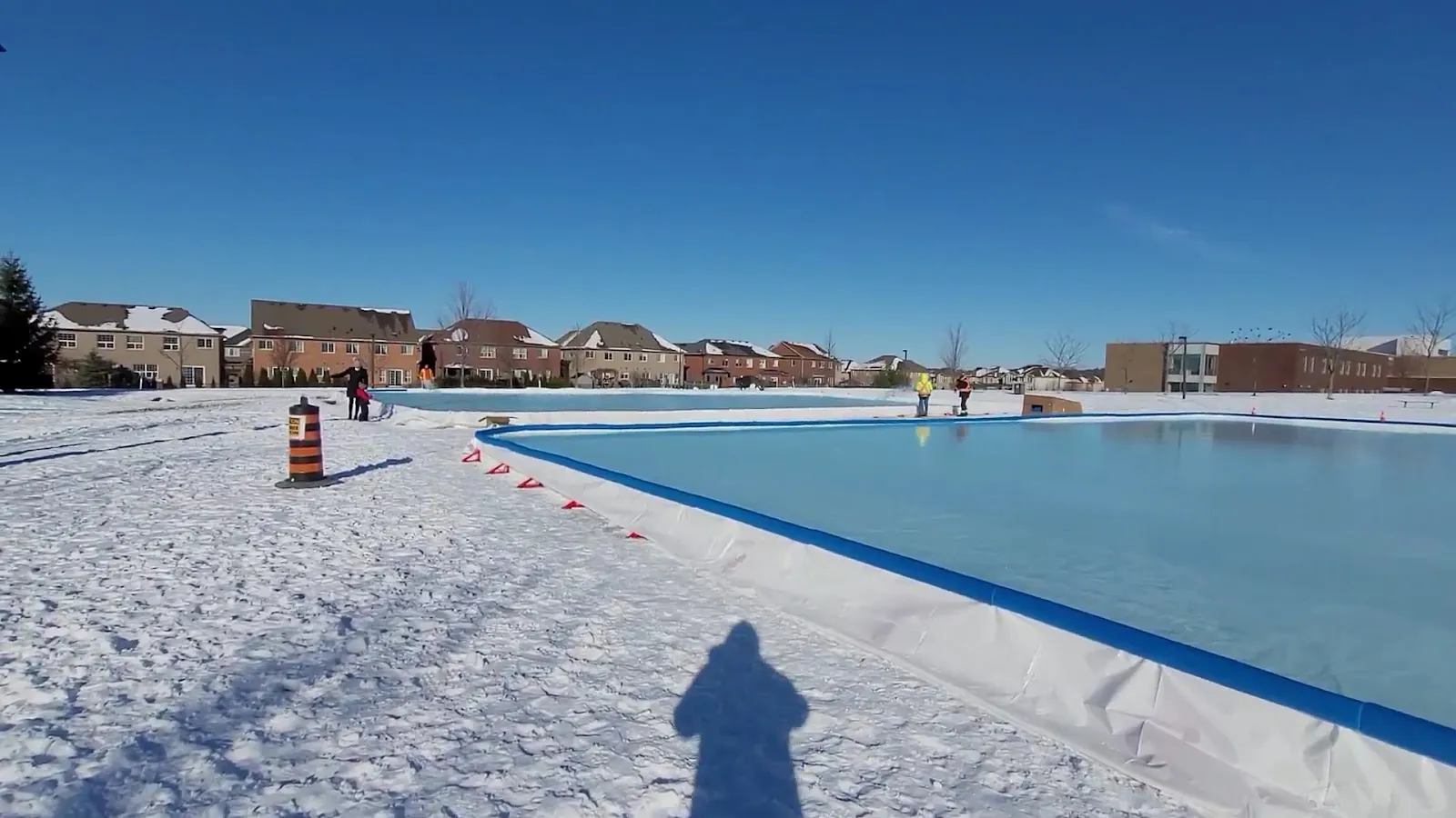 The blue-lined outdoor rink at Ada Johnson Park in Aurora in winter, with snowy grass around the liner and houses beyond — Ada Johnson Park Outdoor Rinks
