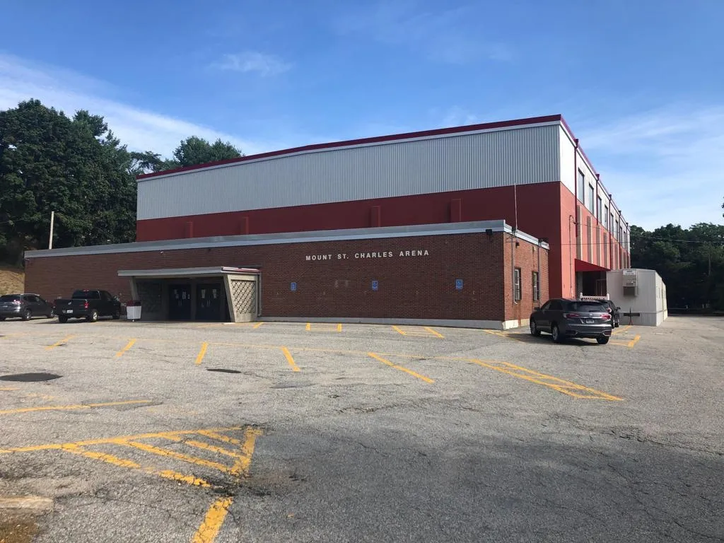 The brick and metal-clad Mount St. Charles Arena seen across its car park, a low entrance block under the lettered name and a pale blue sky behind. — Adelard Arena