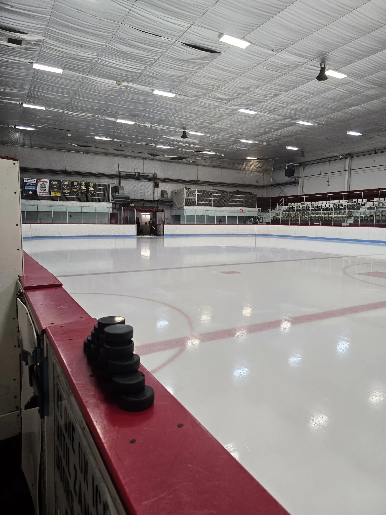 Empty ice sheet seen from rinkside, with hockey pucks stacked on the red dasher boards in the foreground, championship banners on the far wall, and empty bleacher seating. — Adelard Arena