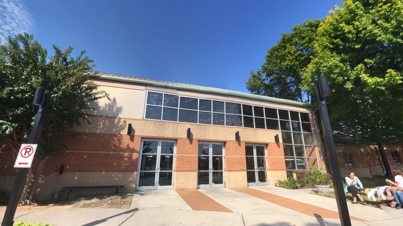 Front of the building on a sunny day: brick and tan facade with tall storefront windows and glass entry doors, trees on either side. — Aerodrome Ice Skating Complex