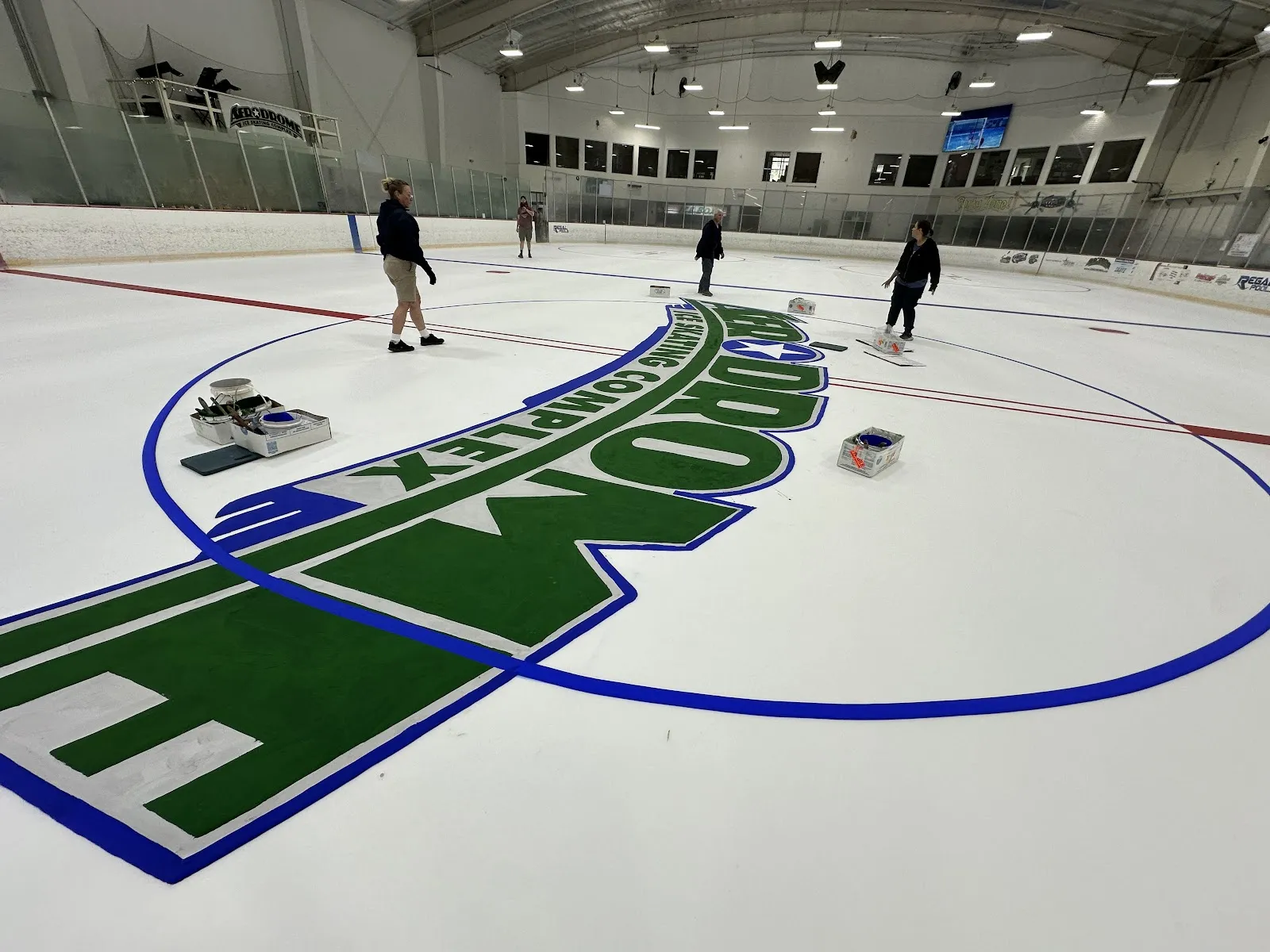 Wide shot of the ice sheet with the 'AERODROME ICE SKATING COMPLEX' logo freshly repainted at center ice; a few staff members are on the ice with paint trays, an 'AERODROME' banner on the mezzanine rail and dasherboard ads along the boards. — Aerodrome Ice Skating Complex