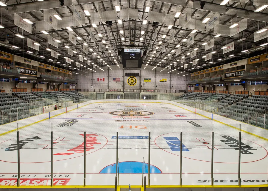 El hielo vacío de Affinity Place desde detrás de la línea de gol: el logotipo de los Bruins en el centro del hielo, asientos grises en ambos lados, letreros de Affinity Place en el nivel de palcos y el marcador sobre el centro. — Affinity Place