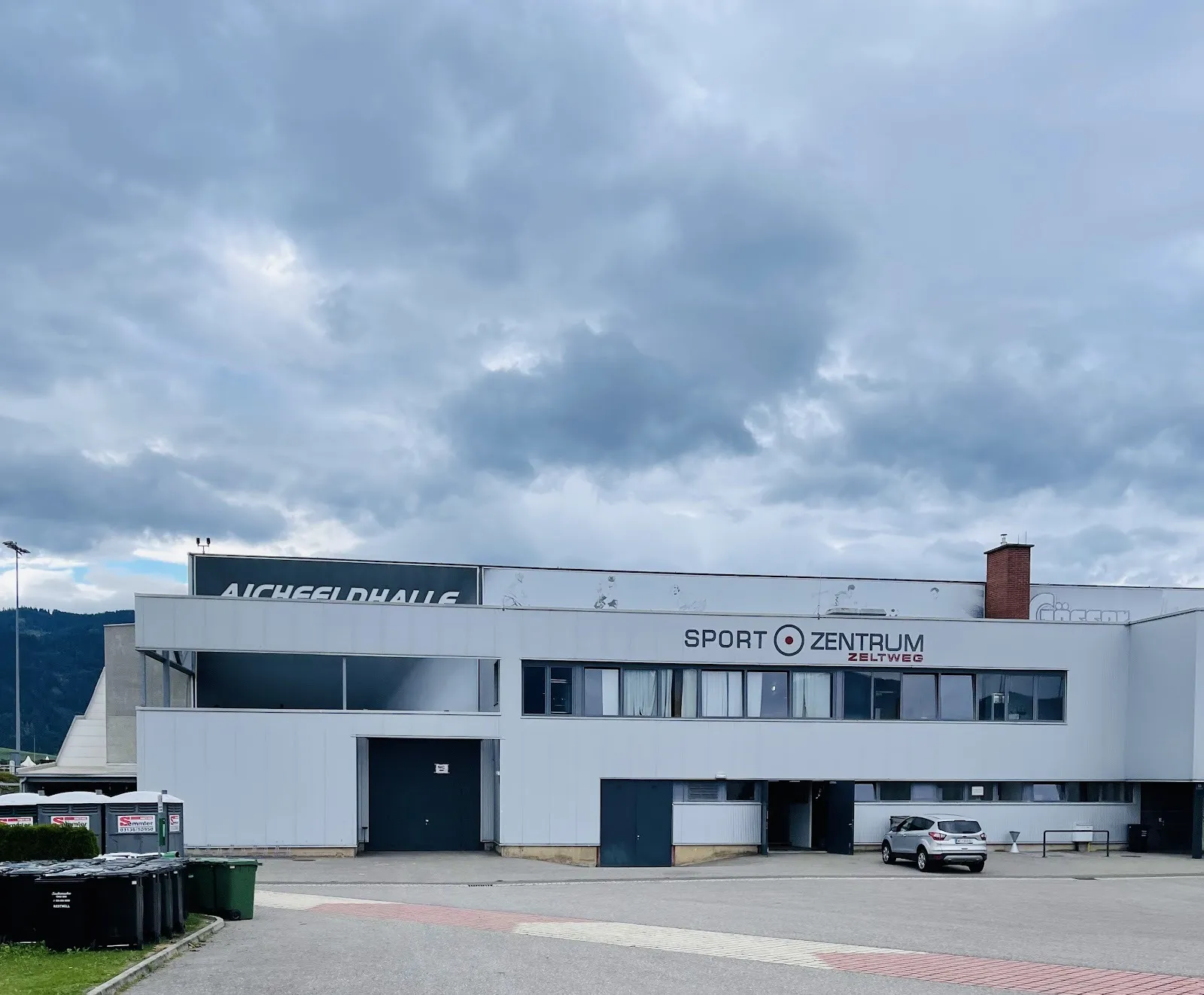 The white and grey Aichfeldhalle sports centre in Zeltweg with its name along the upper wall, seen across the asphalt forecourt under a cloudy sky — Aichfeldhalle Zeltweg