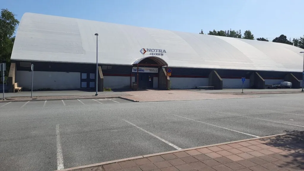 The curved white roof and entrance of Äijänsuon harjoitusjäähalli seen across its car park on a sunny day. – Äijänsuon harjoitusjäähalli