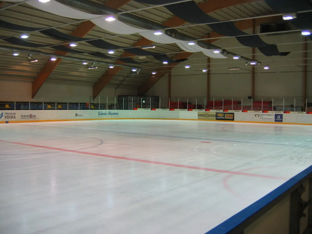 The empty ice sheet of Äijänsuon harjoitusjäähalli with its boards, a timber-framed roof and rows of lights overhead. – Äijänsuon harjoitusjäähalli