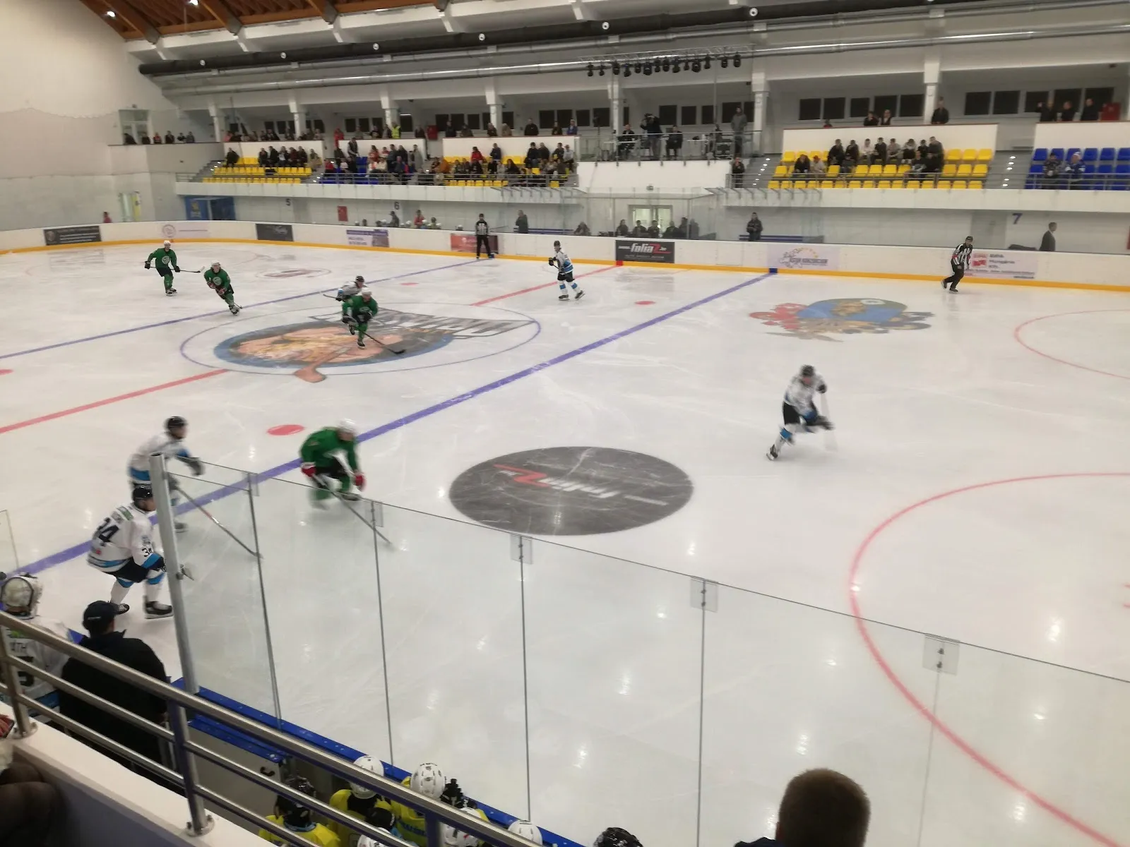 A game on the ice of the Ajka ice hall, seen from the stands behind the glass, with the centre-ice logo and the boards. — Ajka Jégcsarnok