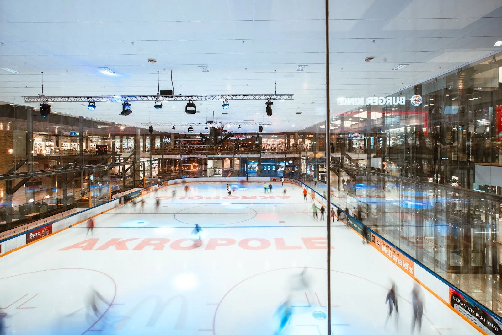 The ice sheet with the AKROPOLE lettering on the ice, seen through the glass from the gallery, with the mall's shop floors behind it. — Akropole Rīga ledus arēna