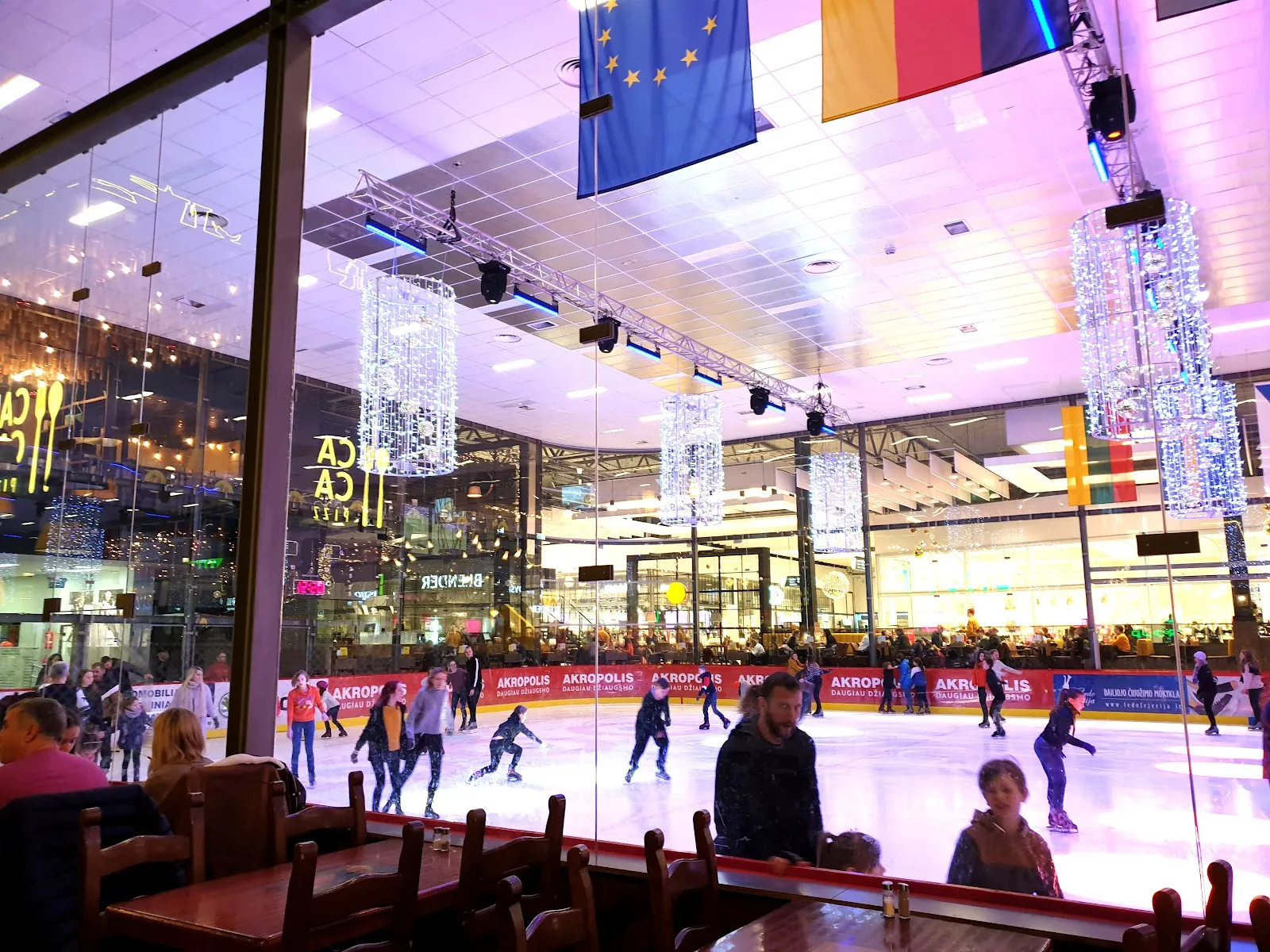 The Akropolis Vilnius ice rink seen through the glass wall during a public session, with skaters across the sheet and national flags hung above the ice. — Akropolio ledas Vilniuje