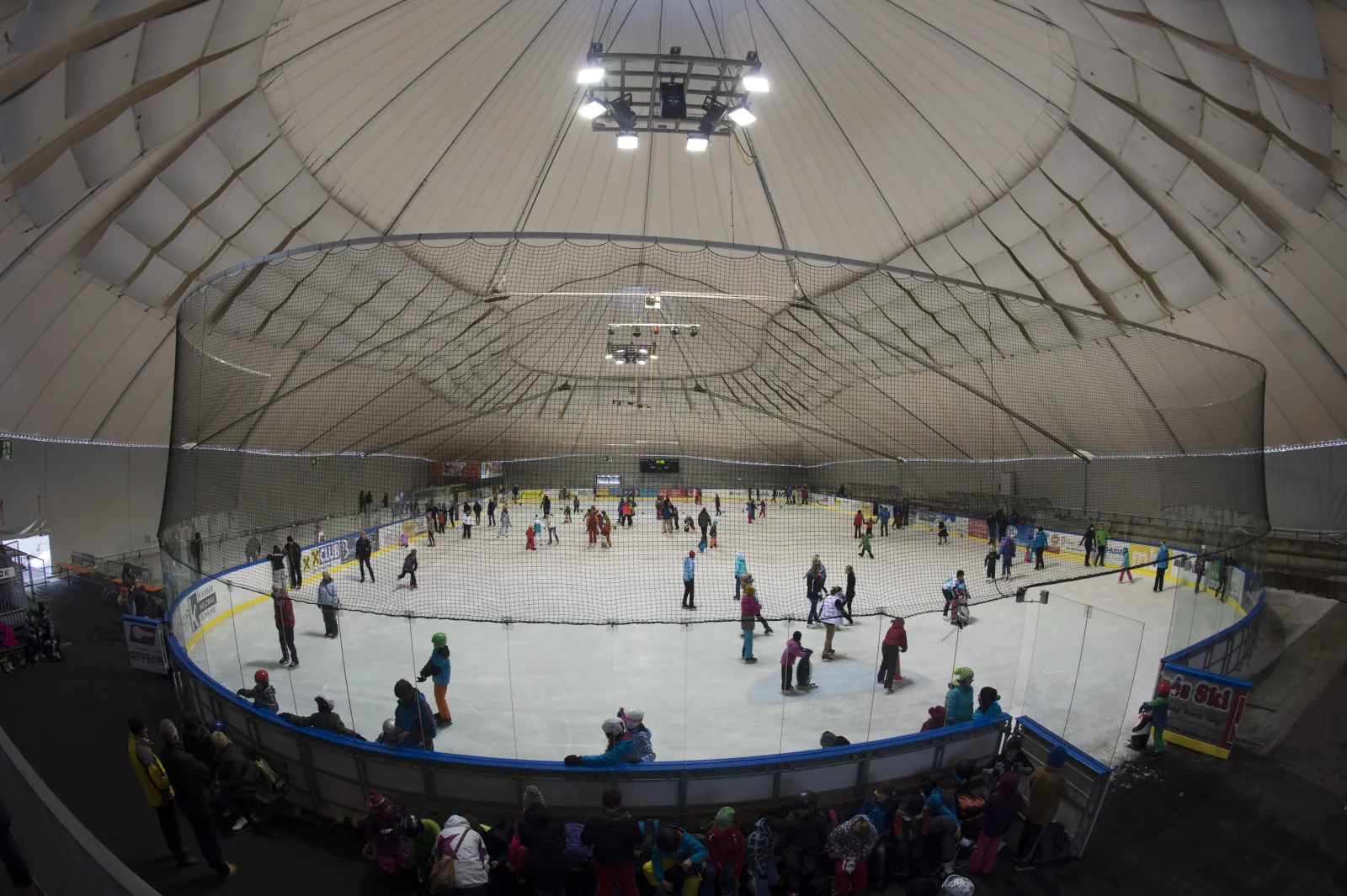 Weitansicht der überdachten Eisbahn im Aktivpark Montafon, Schlittschuhläufer auf dem Eis unter der hohen weißen Kuppel, Zuschauer an den Banden. – Kunsteisbahn Aktivpark Montafon