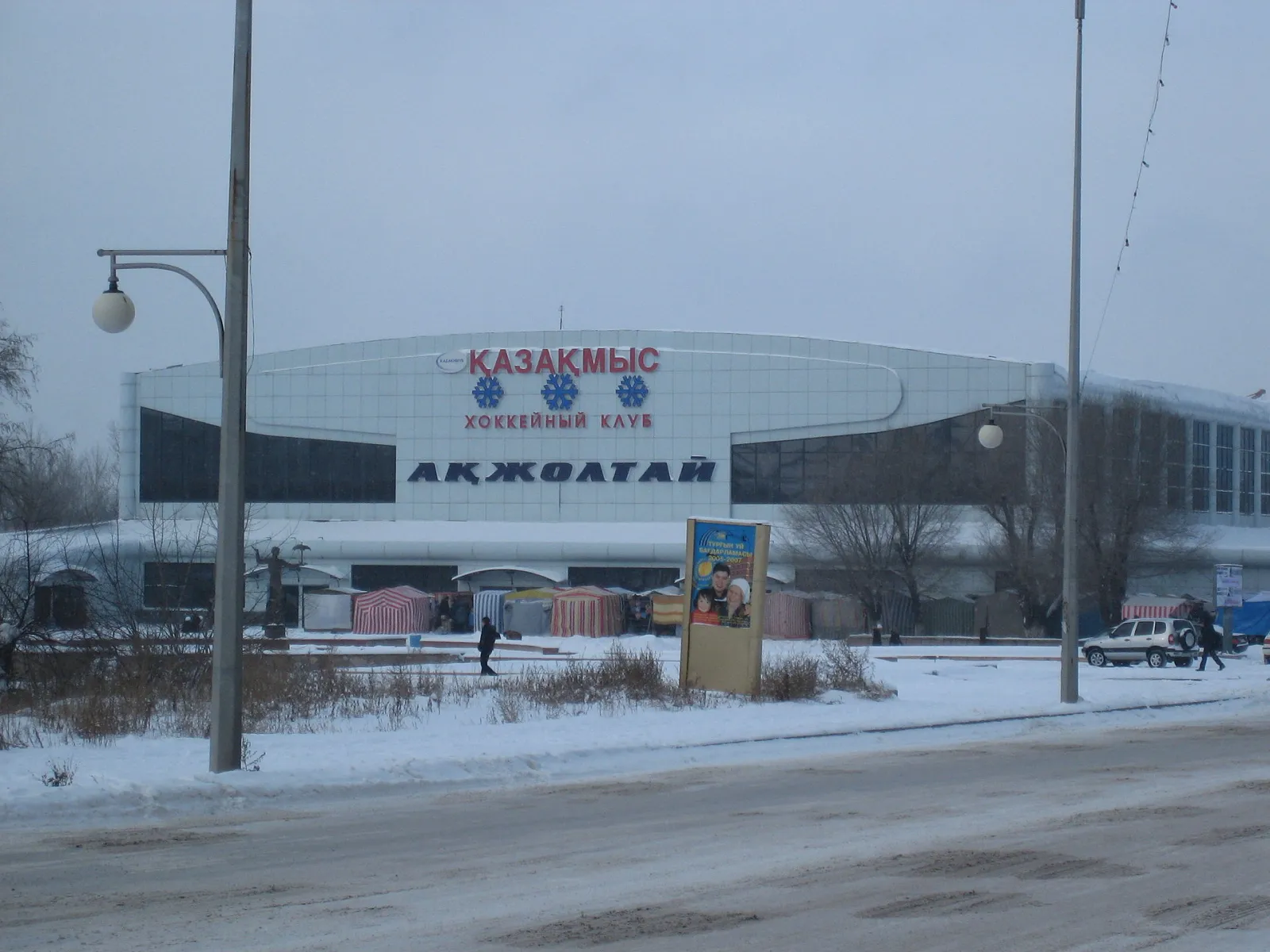 The front of the Akzholtay ice palace in Karaganda, a white-clad hall with the name across the facade, seen from the road in winter — Akzholtay Ice Palace