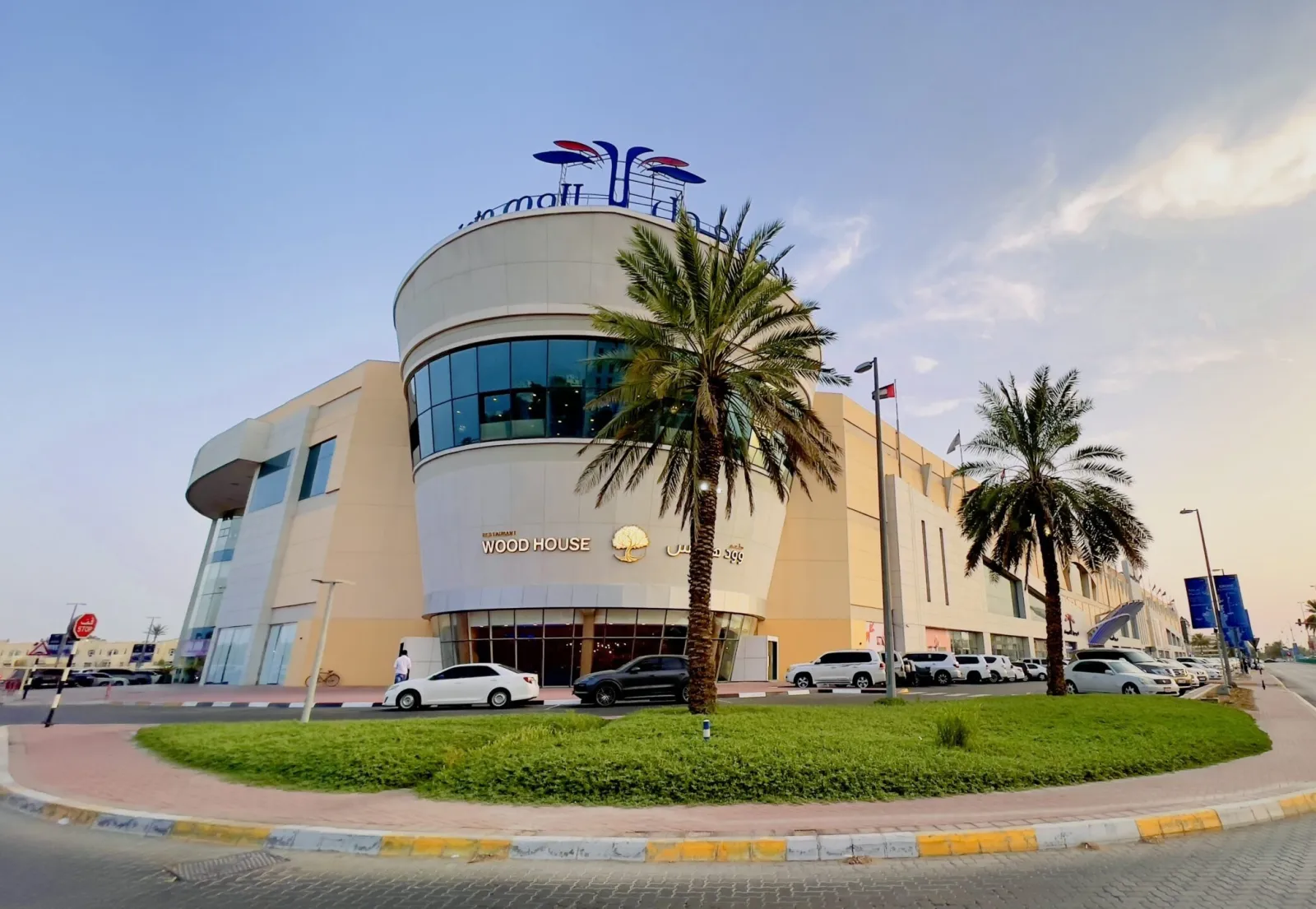 The rounded entrance block of Al Ain Mall with its curved glass front, signage on top and palm trees in the foreground. — Al Ain Mall Ice Rink