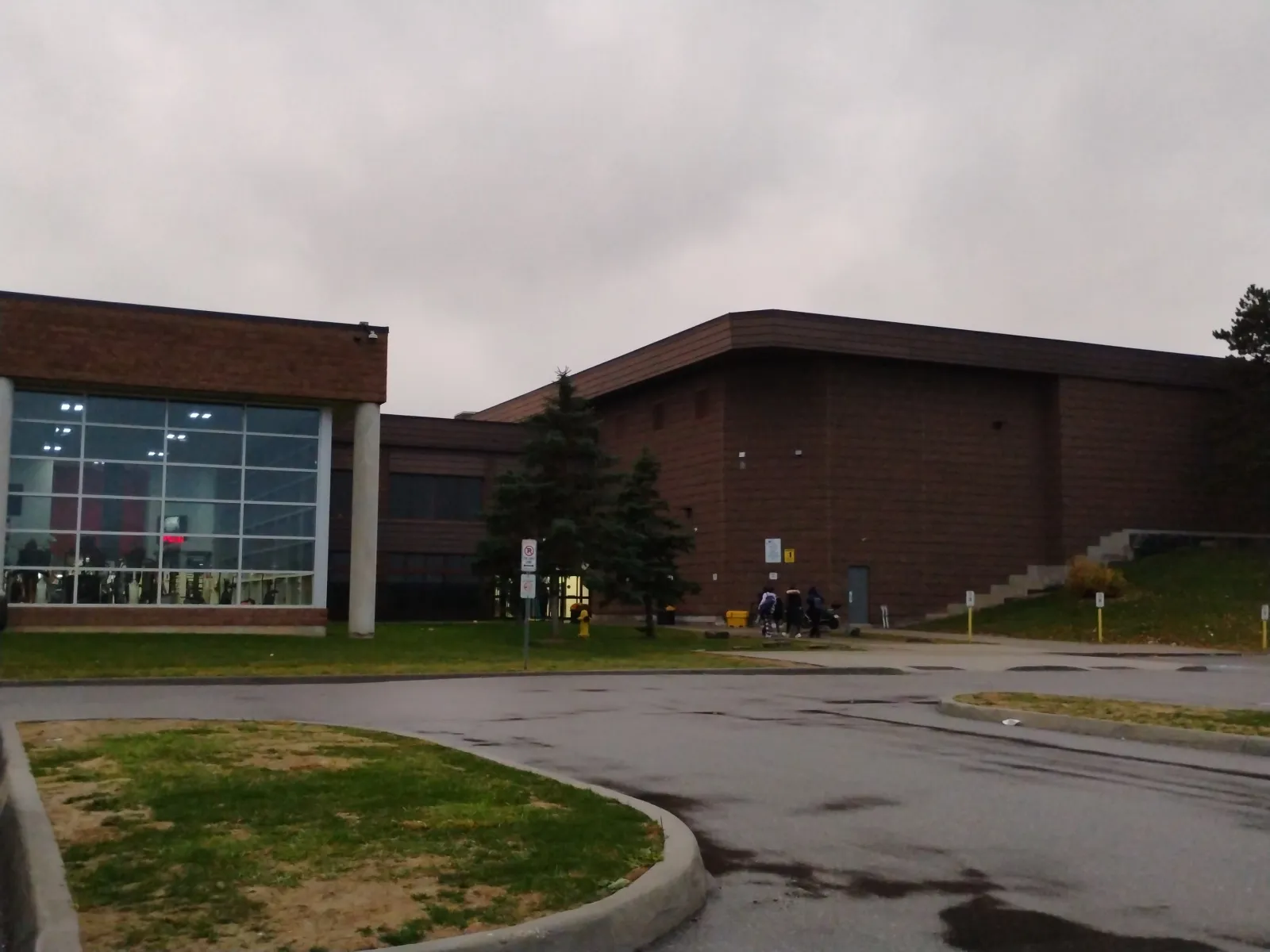 The brown-brick community centre from the parking lot: a glass-walled entrance block on the left and the windowless arena wing behind. — Al Palladini Community Centre (Vaughan)