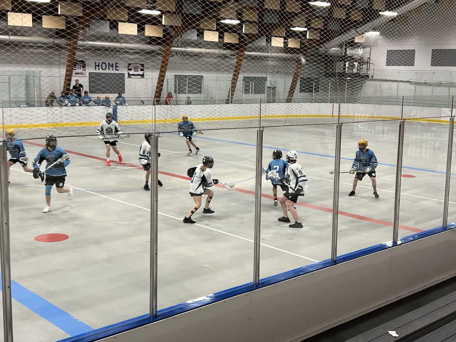 El amplio interior de la arena: toda la superficie de hielo y las vallas, con jugadores de lacrosse o de hockey a media distancia y las gradas detrás. — Al Ritchie Arena