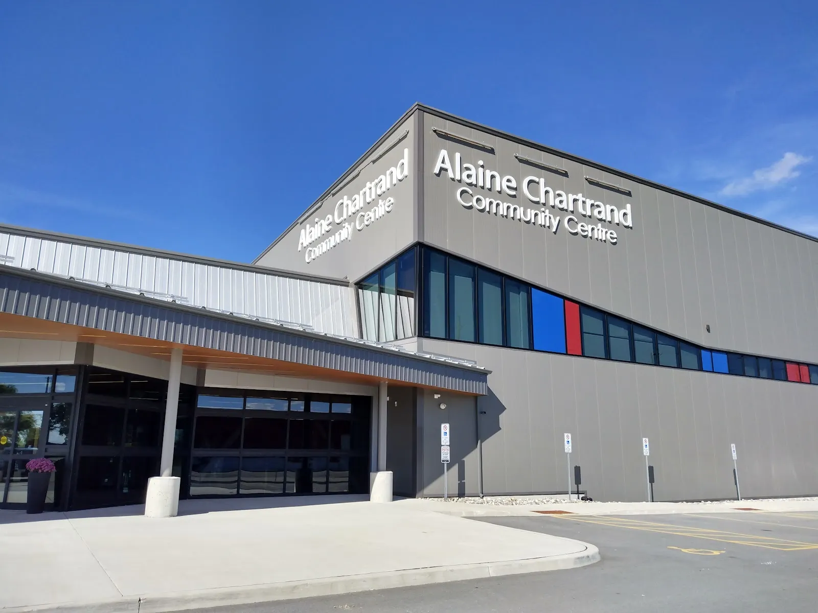 El Alaine Chartrand Community Centre, un edificio revestido de metal gris con su nombre en letras grandes y una franja de ventanas de colores. — Alaine Chartrand Community Centre