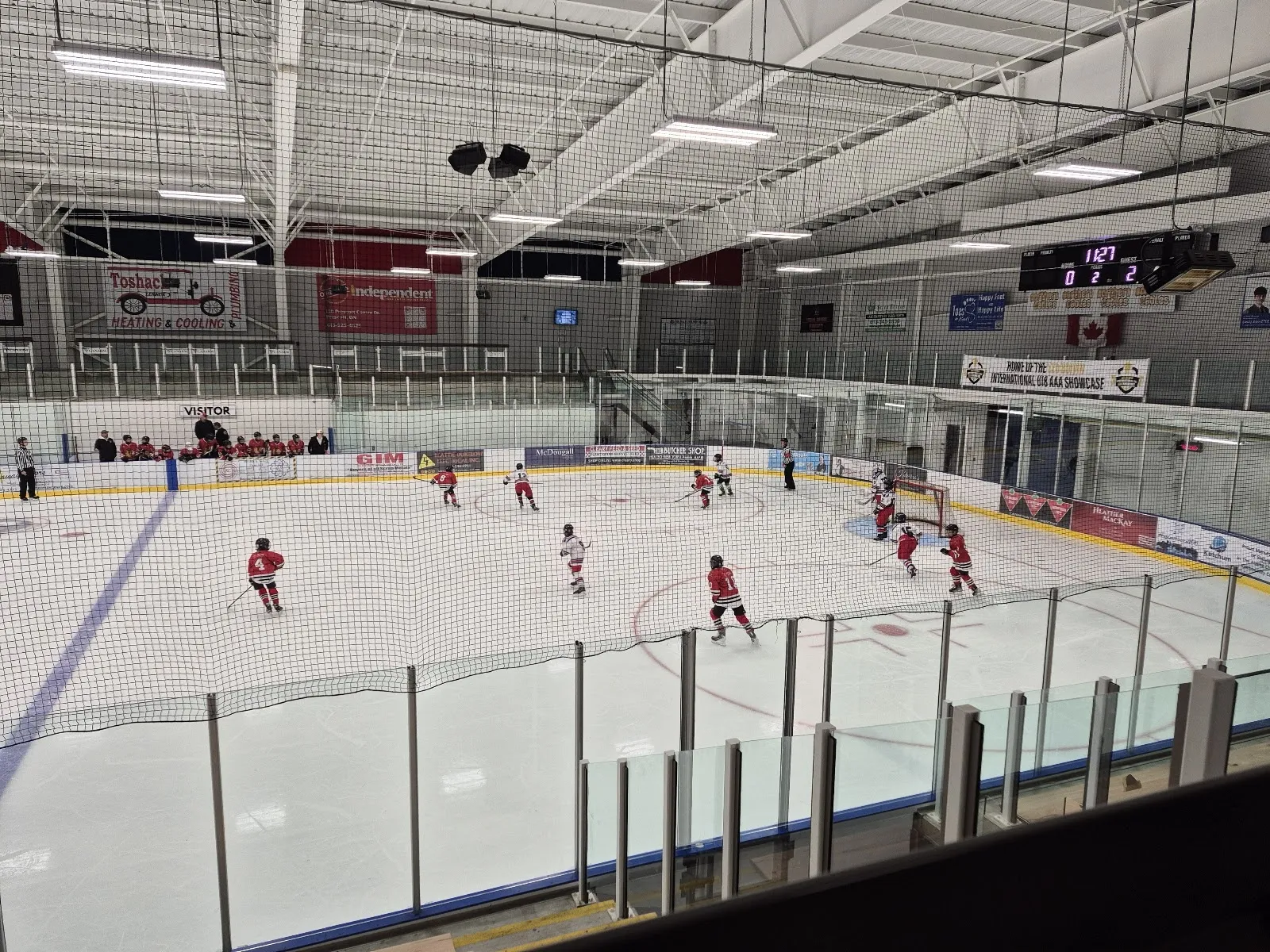 Un partido visto desde el barandal superior a través de una pista amplia, con unos cuantos jugadores de rojo repartidos por el hielo bajo un techo de armaduras de acero. — Alaine Chartrand Community Centre