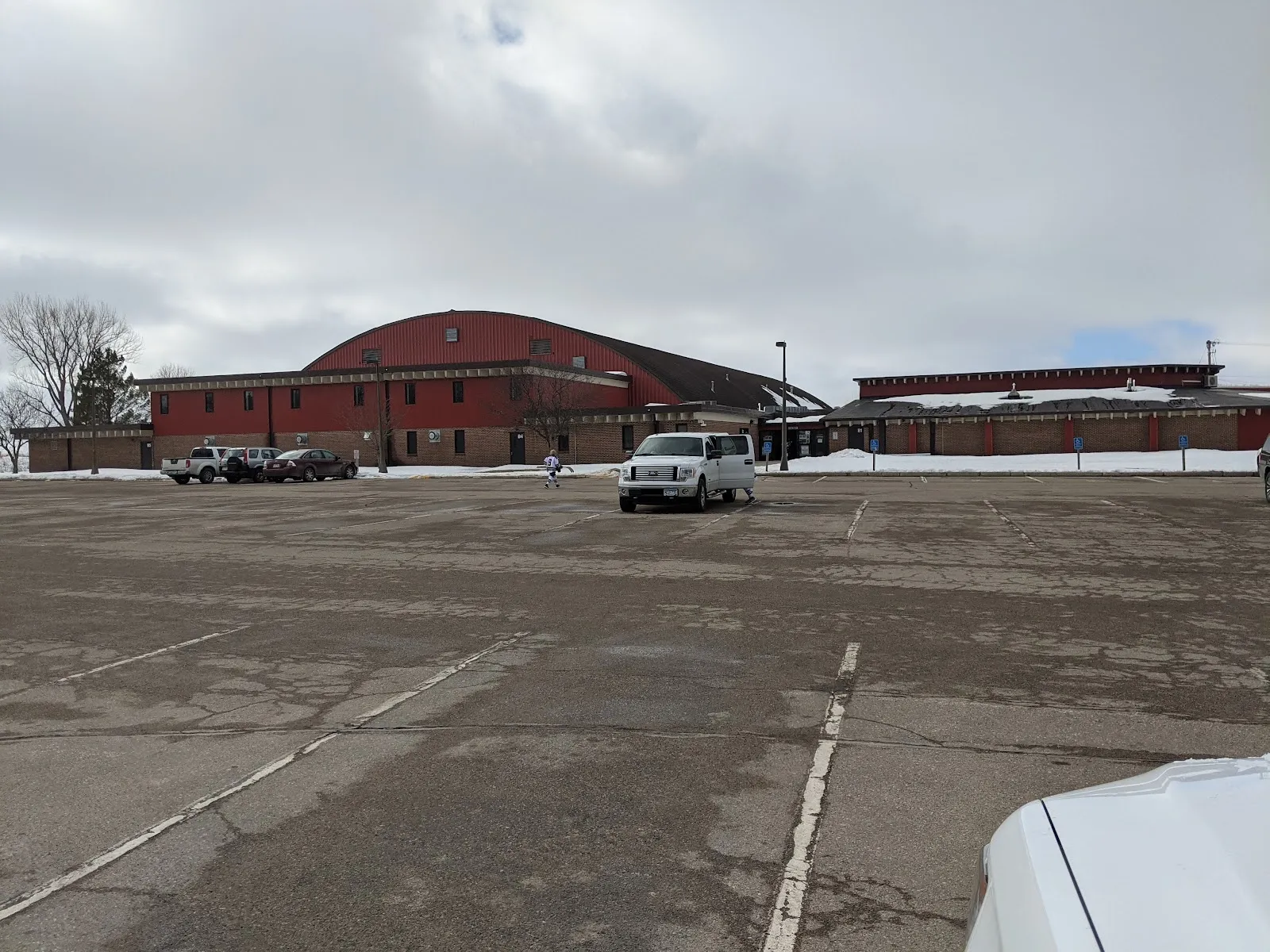 La arena de Albert Lea, de techo rojo abovedado, vista a través de su estacionamiento, con un ala de entrada de ladrillo a la izquierda y árboles sin hojas bajo un cielo gris. — Albert Lea City Arena