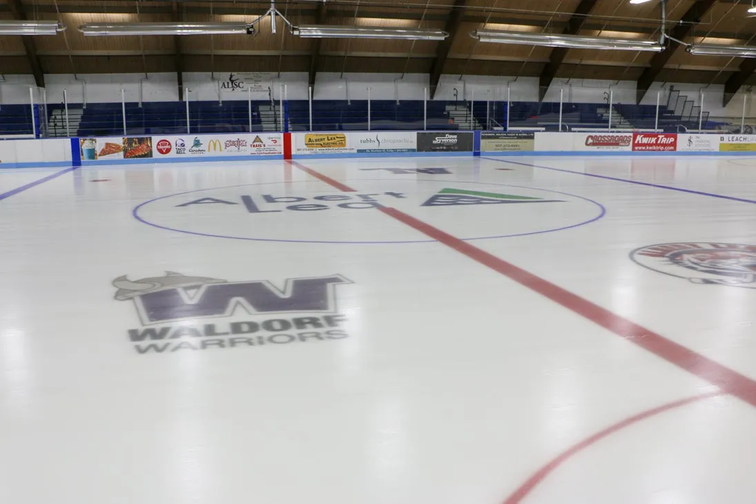 El hielo de la pista Nystrom en el centro, pintado con los logotipos de Albert Lea y de los Waldorf Warriors, con letreros de patrocinadores y asientos azules a lo largo del lado opuesto. — Albert Lea City Arena