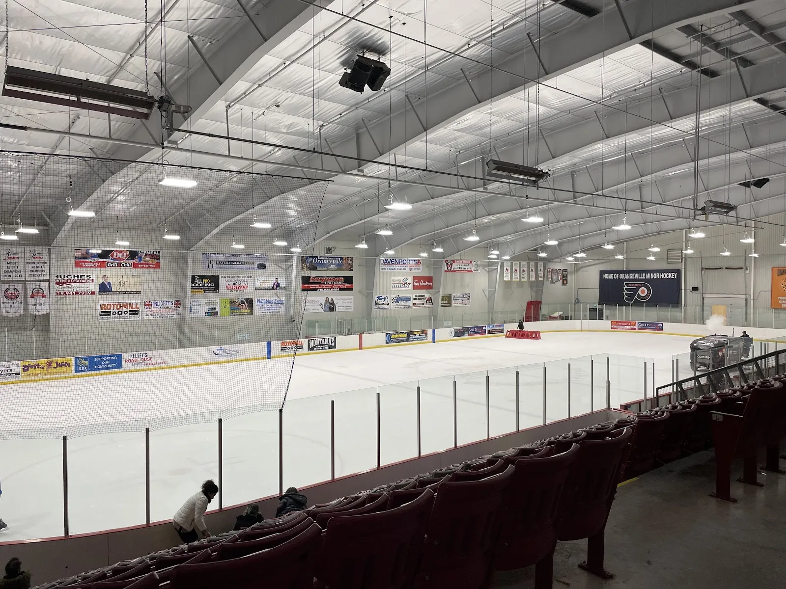 Una pista completa vista desde los asientos rojos bajo un techo blanco curvo, con mantas y letreros de patrocinadores a lo largo de la pared del fondo. — Alder Recreation Centre (Orangeville)