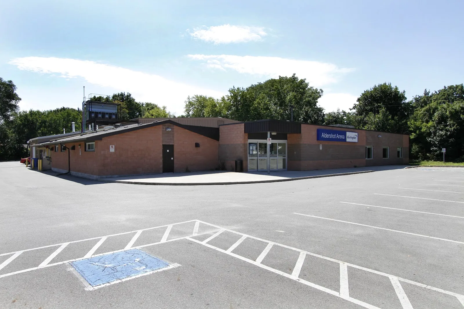 Edificio bajo de arena de ladrillo detrás de un amplio estacionamiento con un espacio de estacionamiento accesible en primer plano y árboles detrás. — Aldershot Arena