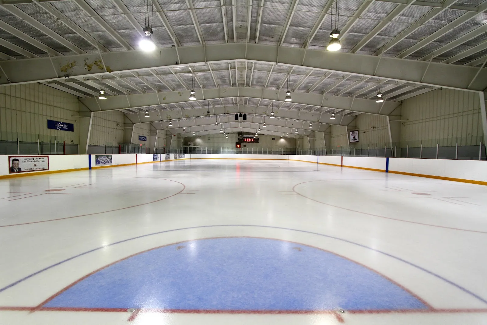 La pista vacía desde las vallas del fondo: hielo recién hecho, área de portería azul y línea roja, con las vallas y las luces hasta el extremo opuesto. — Aldershot Arena