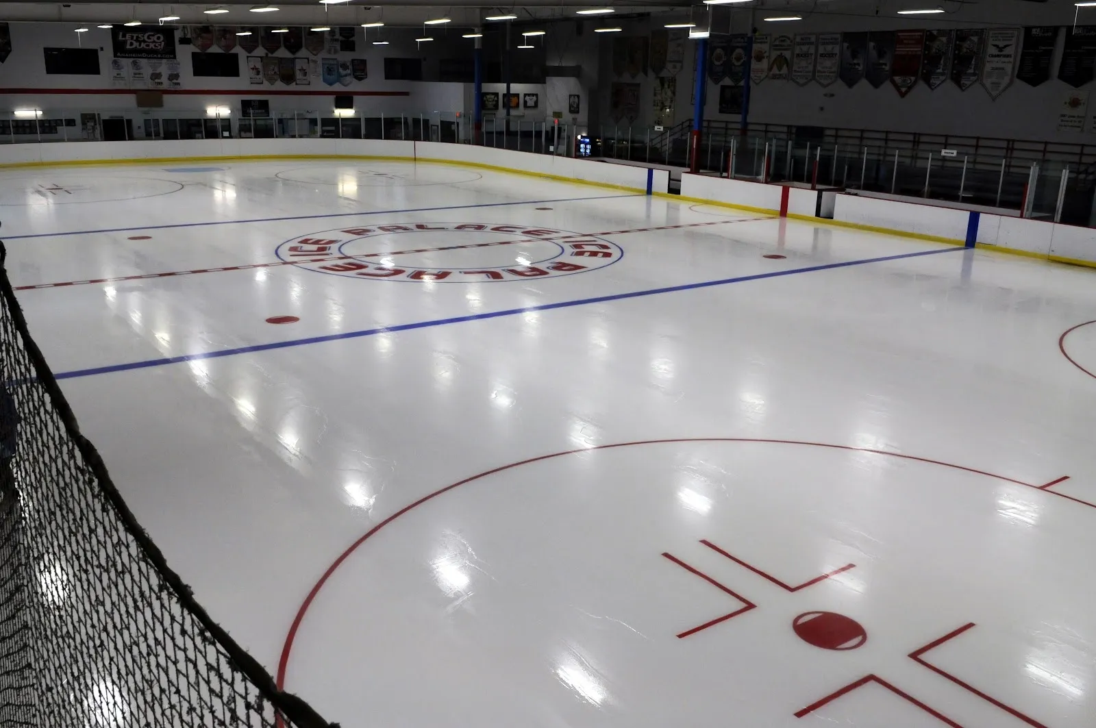 A sheet seen from a corner behind the netting, the red and blue lines and a centre logo on bright white ice under a lit roof. — Aliso Viejo Ice Palace