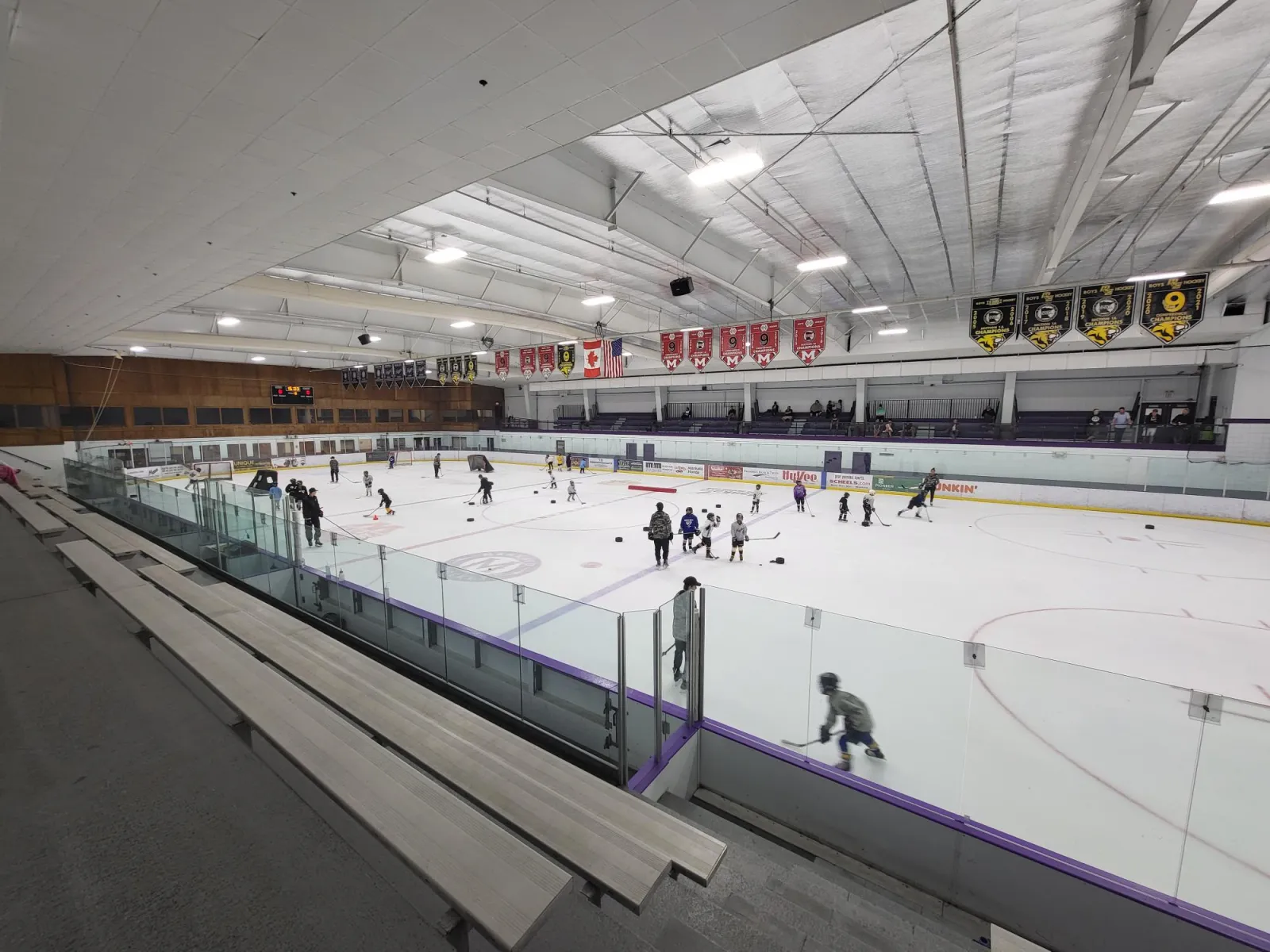 Wide interior view of the rink from the stands during a youth hockey practice, showing the full ice sheet, glass boards, and championship banners hanging from the ceiling. — All Seasons Arena