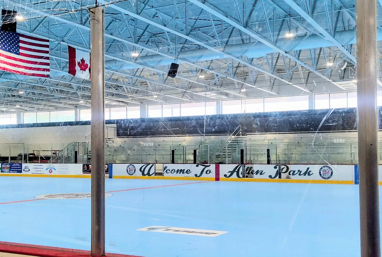 The empty ice sheet seen past two steel columns, with 'Welcome To Allen Park' painted on the boards and US and Canadian flags under the high steel roof. — Frank J. Lada Civic Arena
