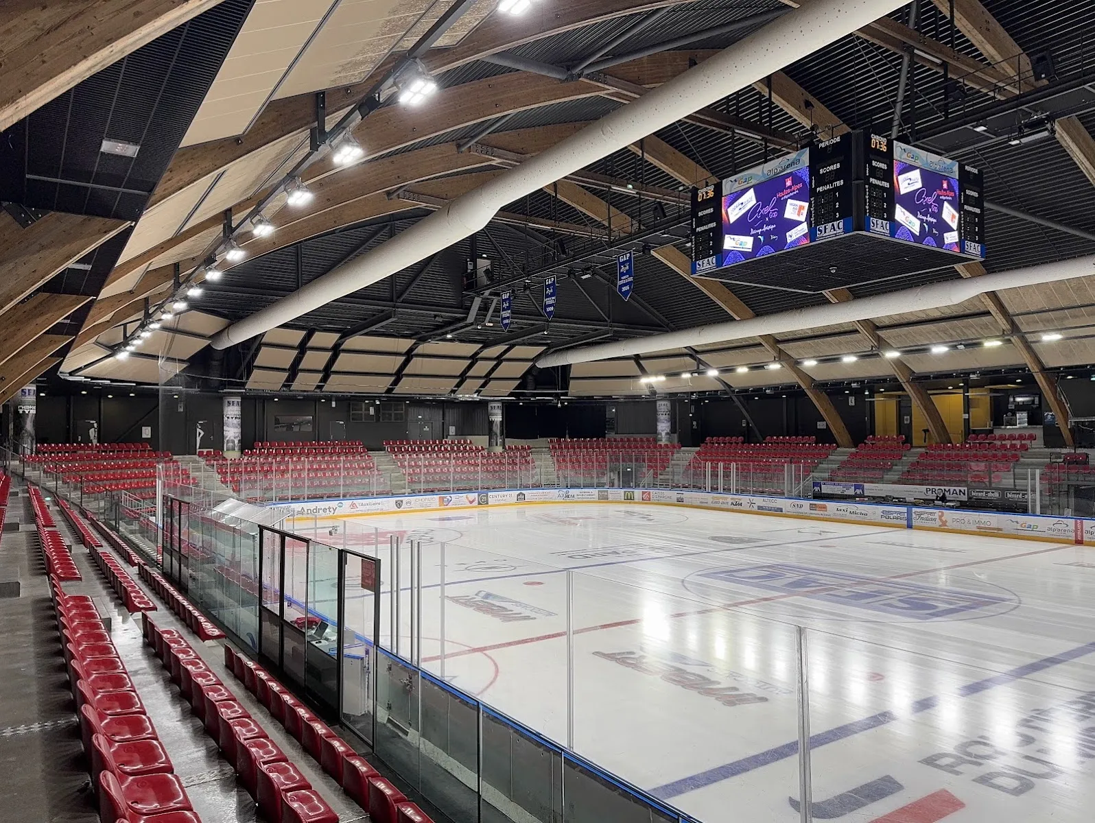 The empty ice and the red stands of the Alp'Arena, seen from the glass with the timber roof arching overhead and the scoreboard above centre ice. — Alp'Arena