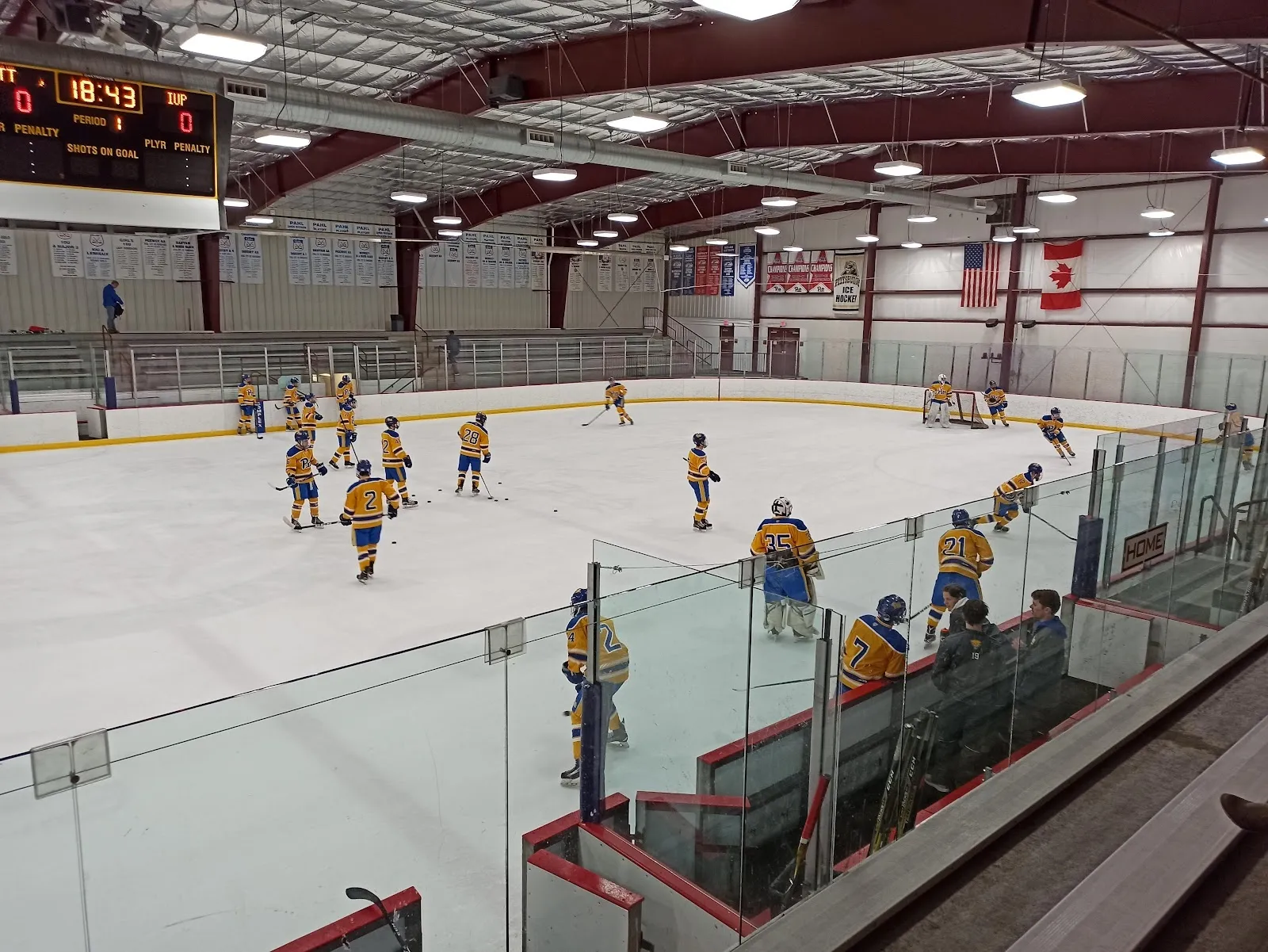 A game in play across a wide sheet seen from the glass, players spread across the ice under a red-trussed roof with a scoreboard on the far wall. — Alpha Ice Complex