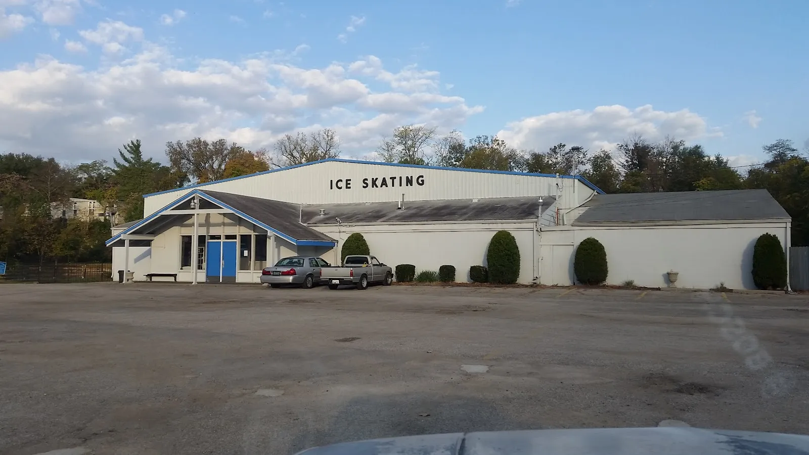 Vista de día del edificio blanco y bajo con una orilla de techo de borde azul que dice "ICE SKATING" en letras grandes, una entrada a dos aguas con puertas azules y un pequeño estacionamiento al frente. — Alpine Ice Arena