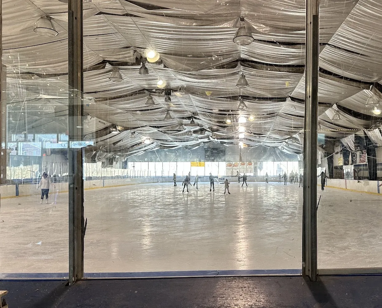 Patinaje público en Alpine Ice Arena visto a través del vidrio del vestíbulo, con patinadores repartidos por el hielo bajo un techo cubierto de telas blancas — Alpine Ice Arena