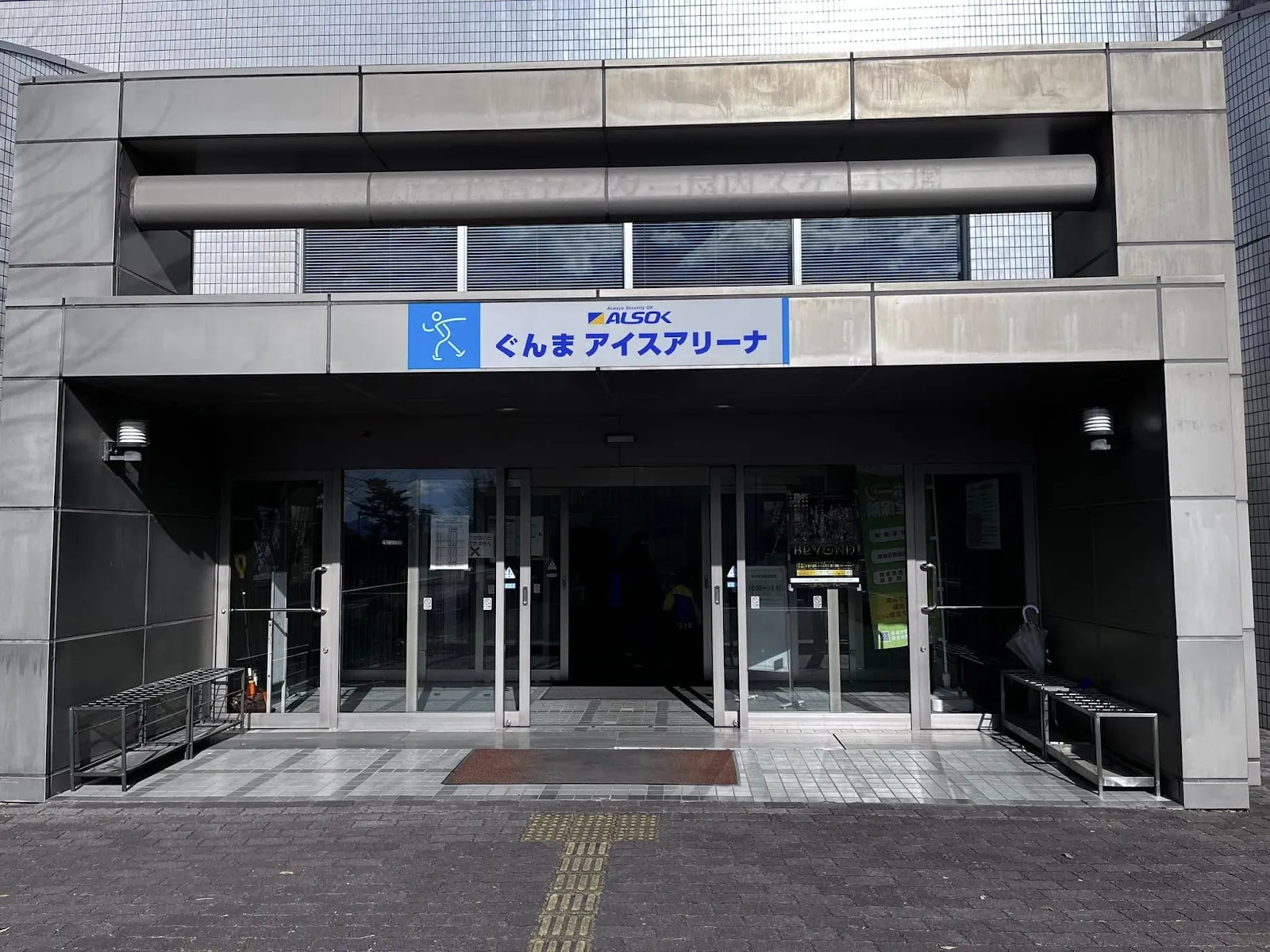 The entrance of ALSOK Gunma Ice Arena in Maebashi, with the blue ALSOK Gunma Ice Arena sign above the glass doors and a ramp at the left — ALSOK Gunma Ice Arena