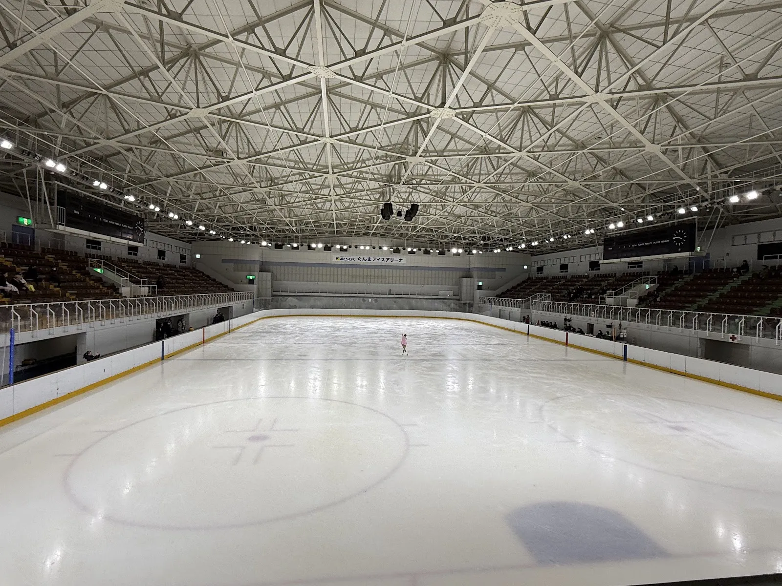 The ice sheet of ALSOK Gunma Ice Arena seen from the boards, with steel roof trusses above, seating along both sides and one skater far down the ice — ALSOK Gunma Ice Arena