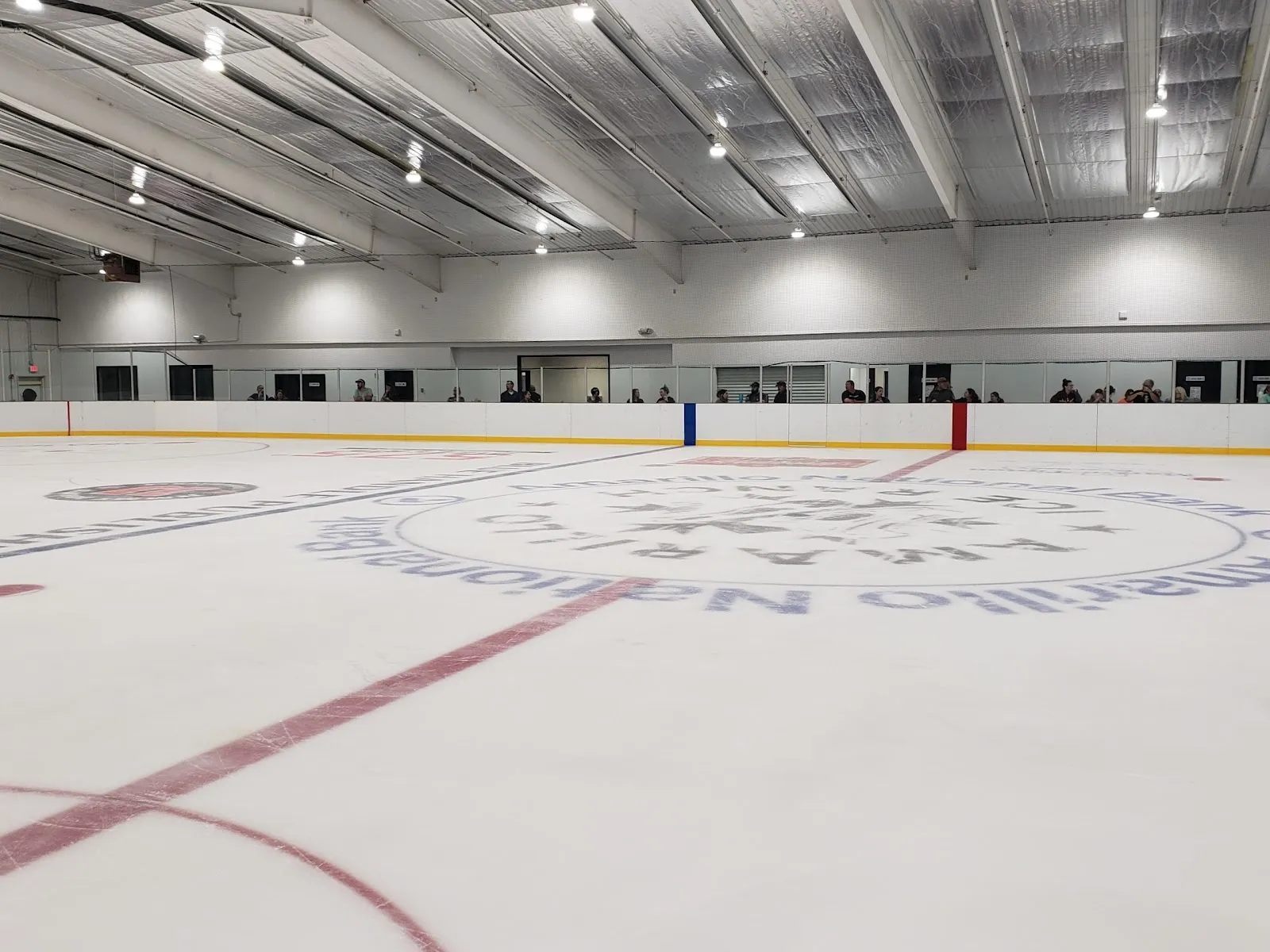 Una pista de hielo vacía bajo un techo aislado plateado, con un logotipo de un patrocinador de Amarillo pintado alrededor del centro de la pista y algunos espectadores detrás del cristal en el lado opuesto. — Amarillo Ice Ranch